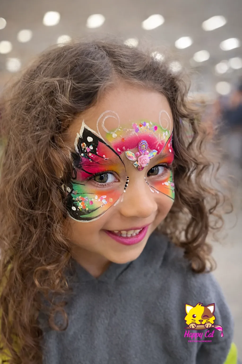 A young girl with curly hair has a colorful butterfly face paint with pink, green, and black shades, white dots, and glitter. She is smiling with her teeth visible and wearing a gray hoodie. There is a Happy Cat logo in the bottom right corner.