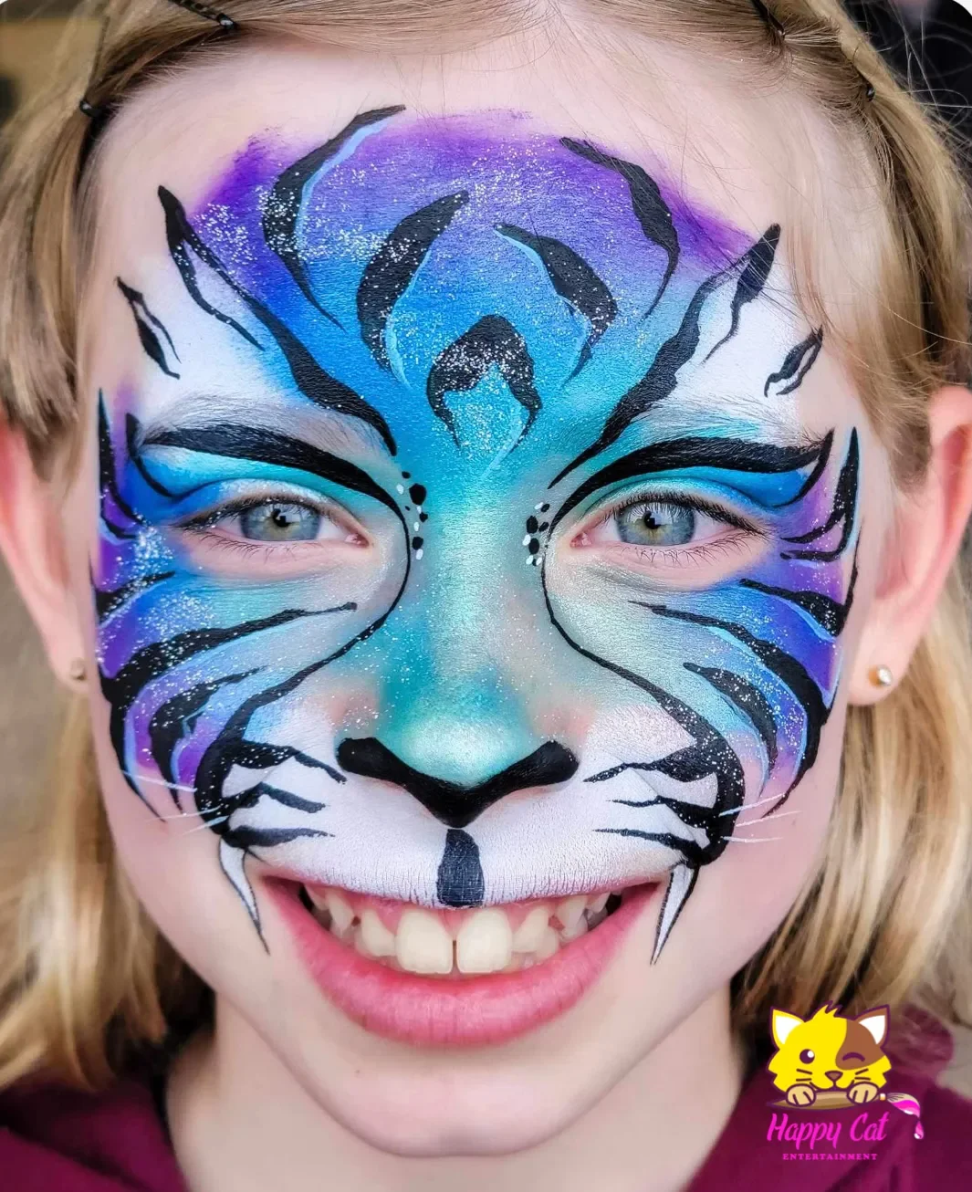 A person with detailed tiger face paint in bright colors, including blue, purple, white, and black, with a Pixar-style cartoon logo of a happy cat in the bottom right corner.