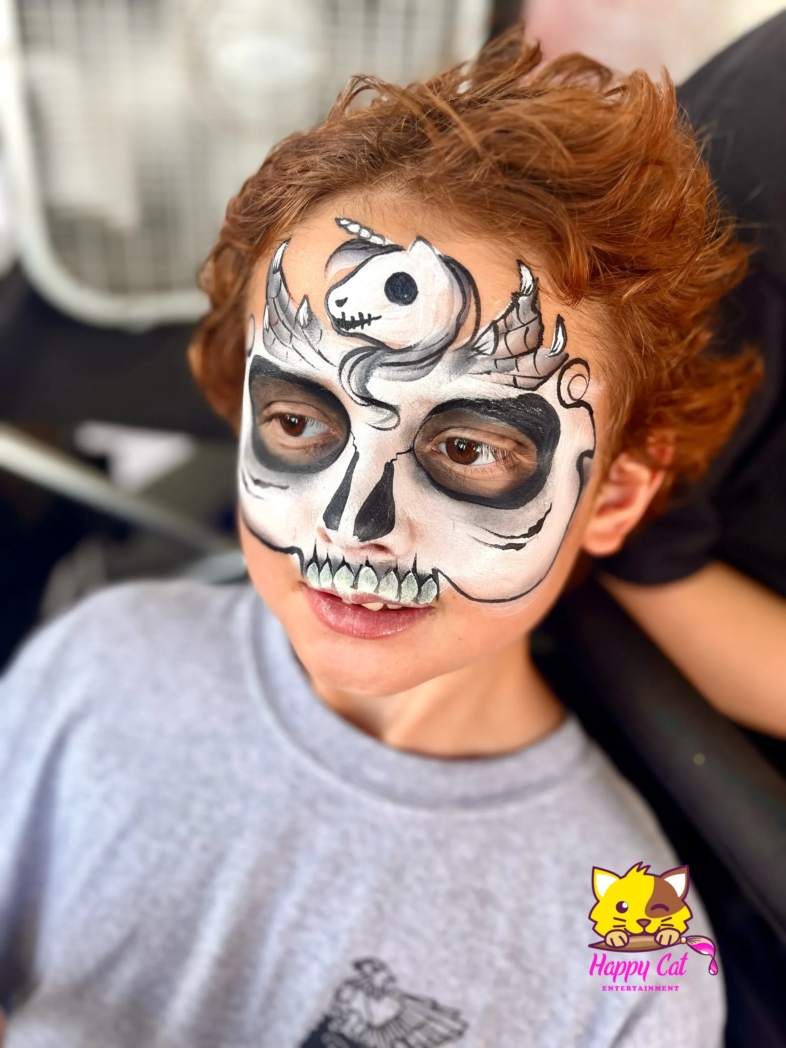 Child with face painted as a skeleton with a unicorn skull, dark eye sockets, and teeth, wearing a gray shirt, smiling.