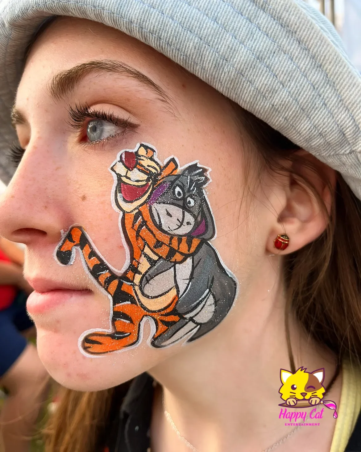 A young girl with painted face of Tigger and Eeyore from Winnie the Pooh, Wearing a light gray hat and a red and gold elf earring.