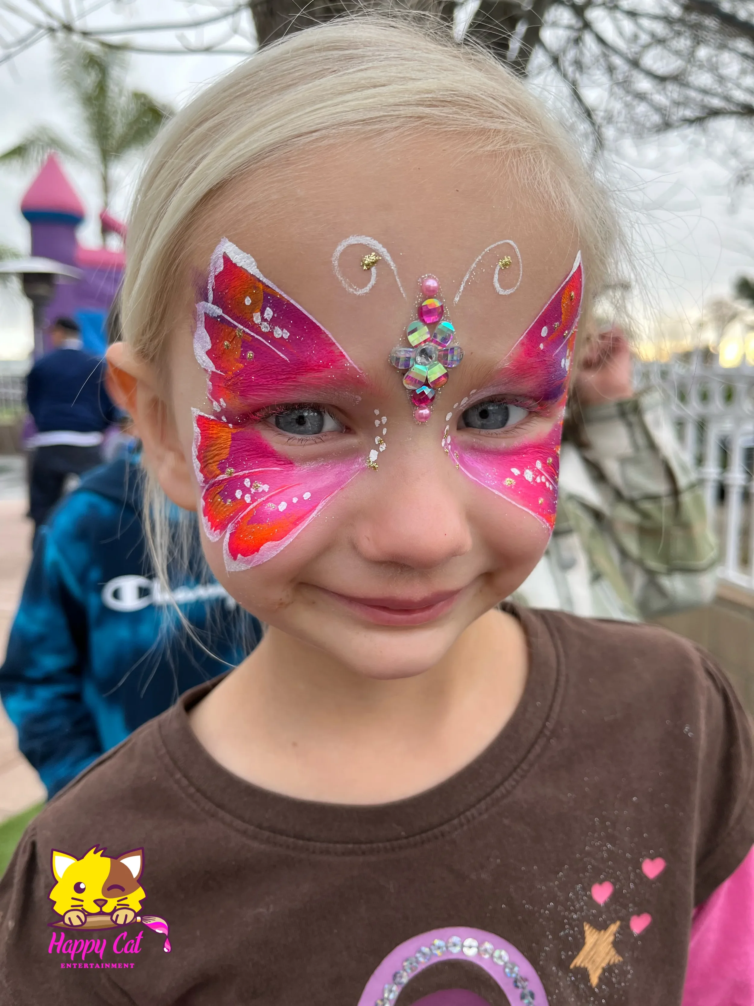 A young girl with blonde hair and blue eyes smiling at the camera. Her face is decorated with pink and orange butterfly face paint and rhinestone embellishments on her forehead. In the background, there are other children and a colorful playground st