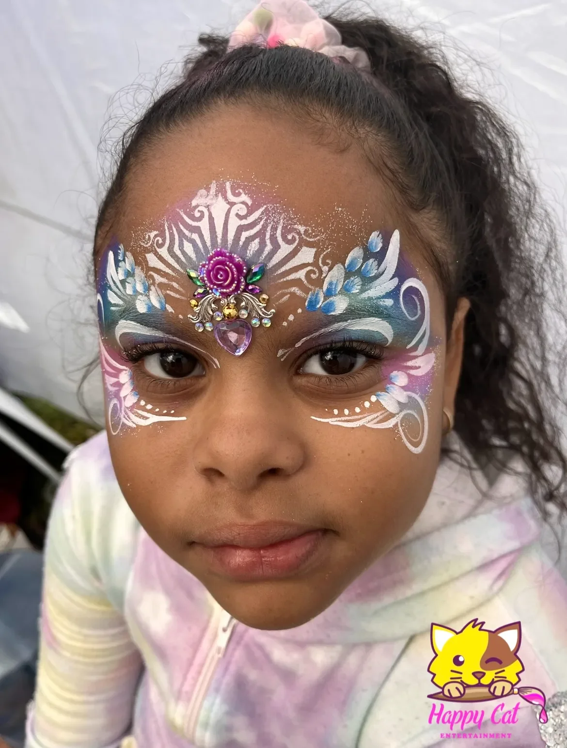 A young girl with intricate face paint in white, blue, purple, and pink with a decorative pink and purple 3D rose embellishment in the center of her forehead, and a small pink heart-shaped gem underneath. She has curly hair tied up with a pink scrunc