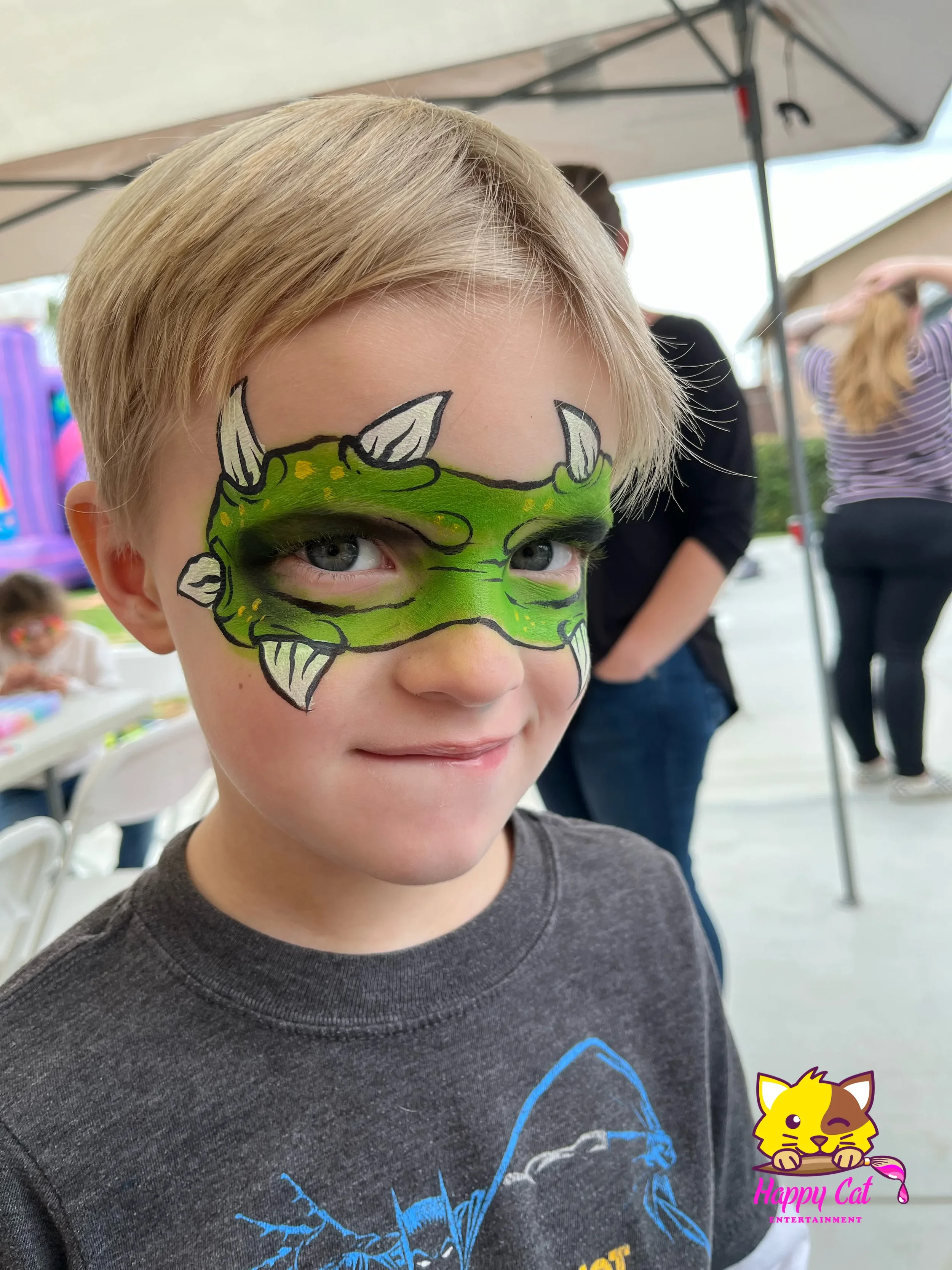 Young boy with face paint of a green dragon with white horns, black outlines, and yellow accents, smiling at an outdoor event under a tent.