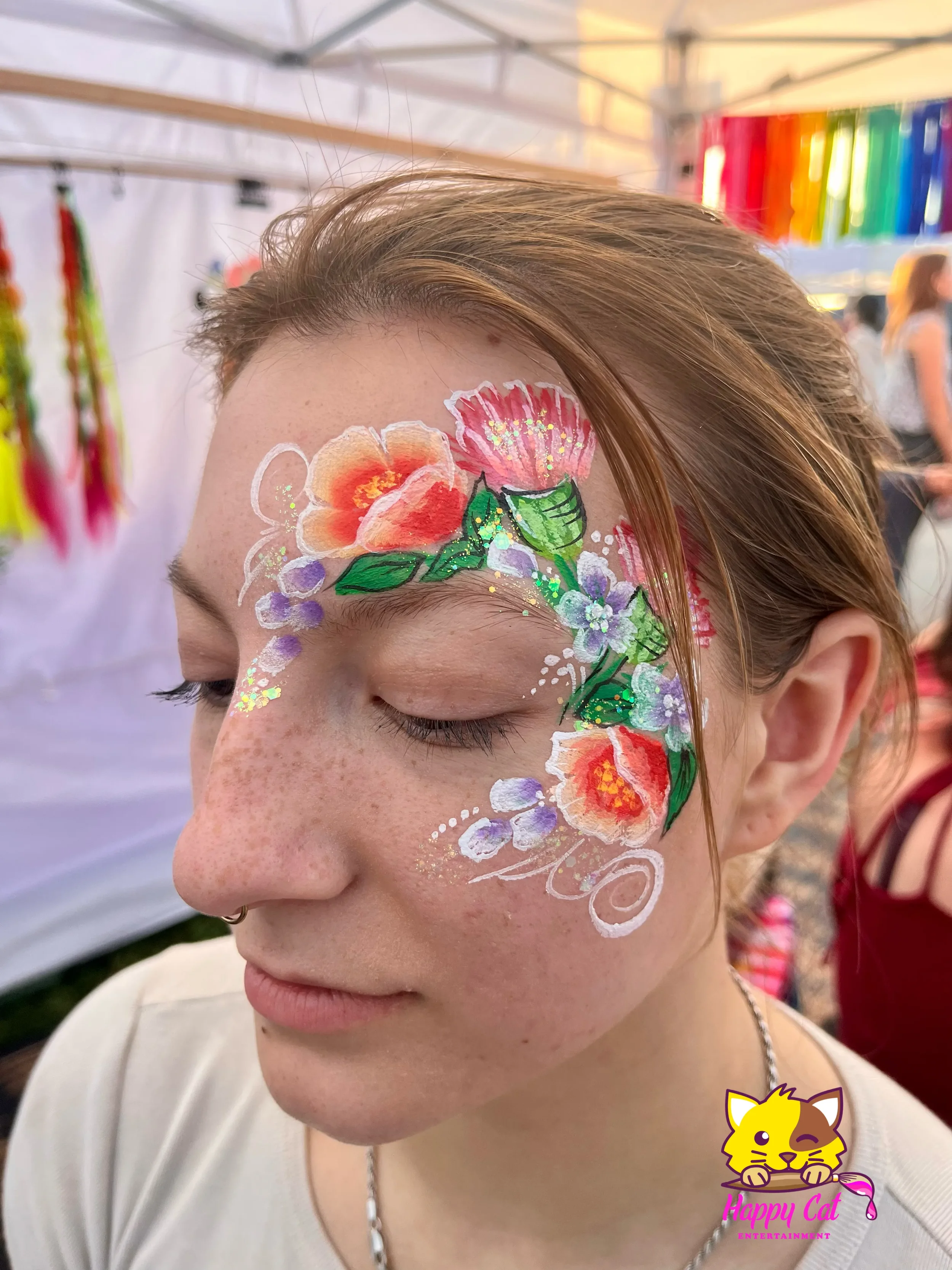 Close-up of a young woman with face paint featuring colorful flowers and leaves, with her eyes closed at an outdoor event.