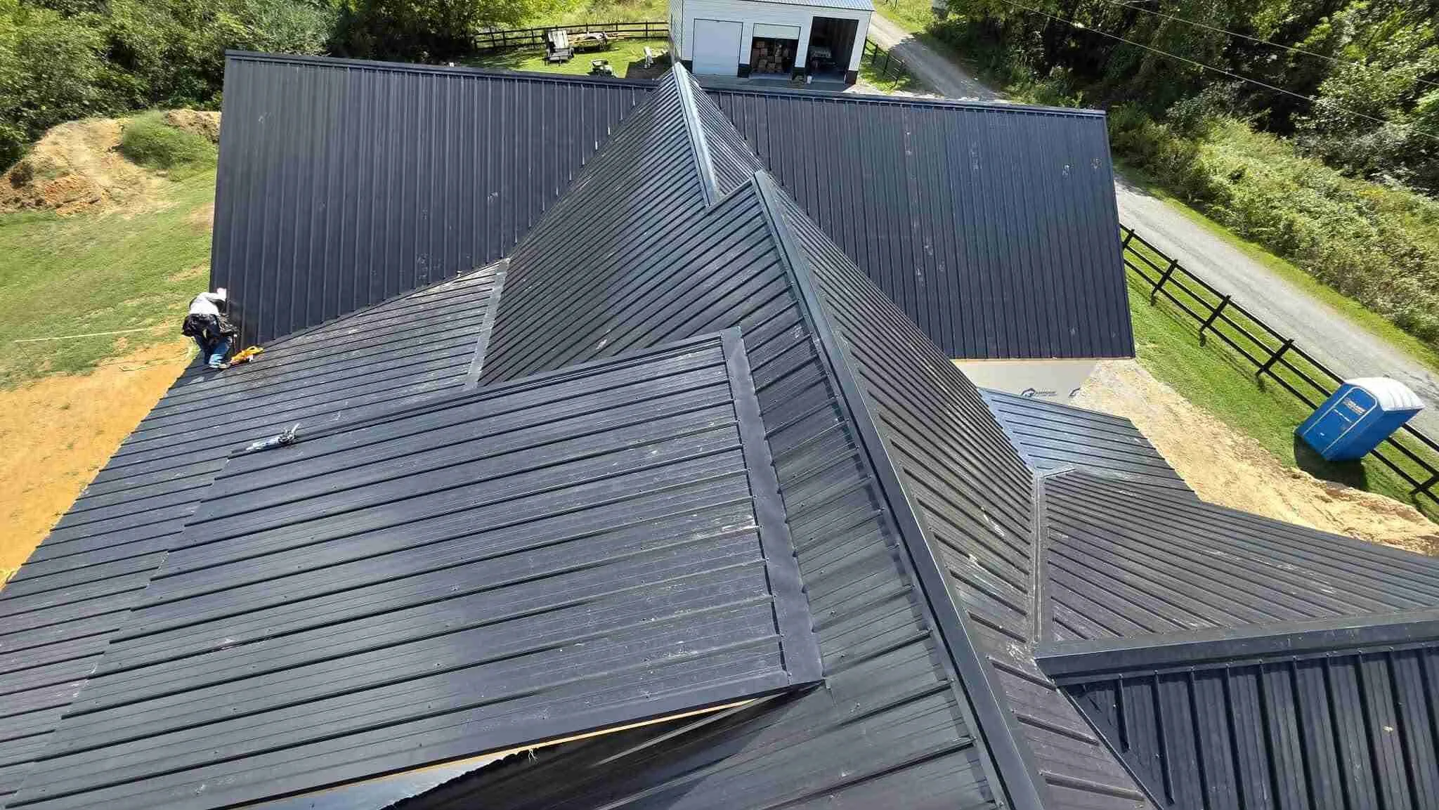 An aerial view of a house roof under construction, with a worker installing or inspecting the black metal roofing panels.