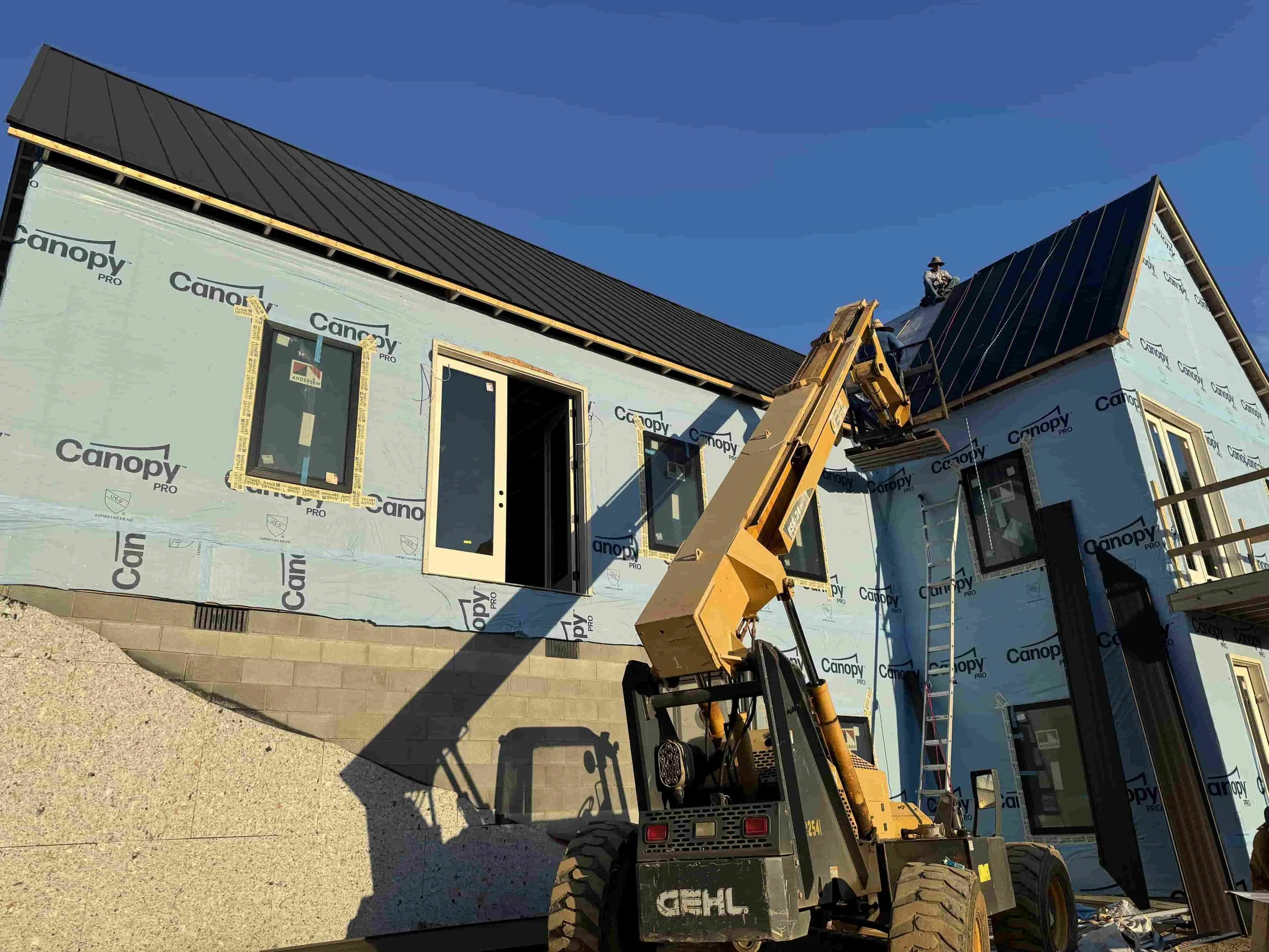 Construction workers installing a black metal roof on a new house, with windows and a ladder, using a yellow telescopic forklift.