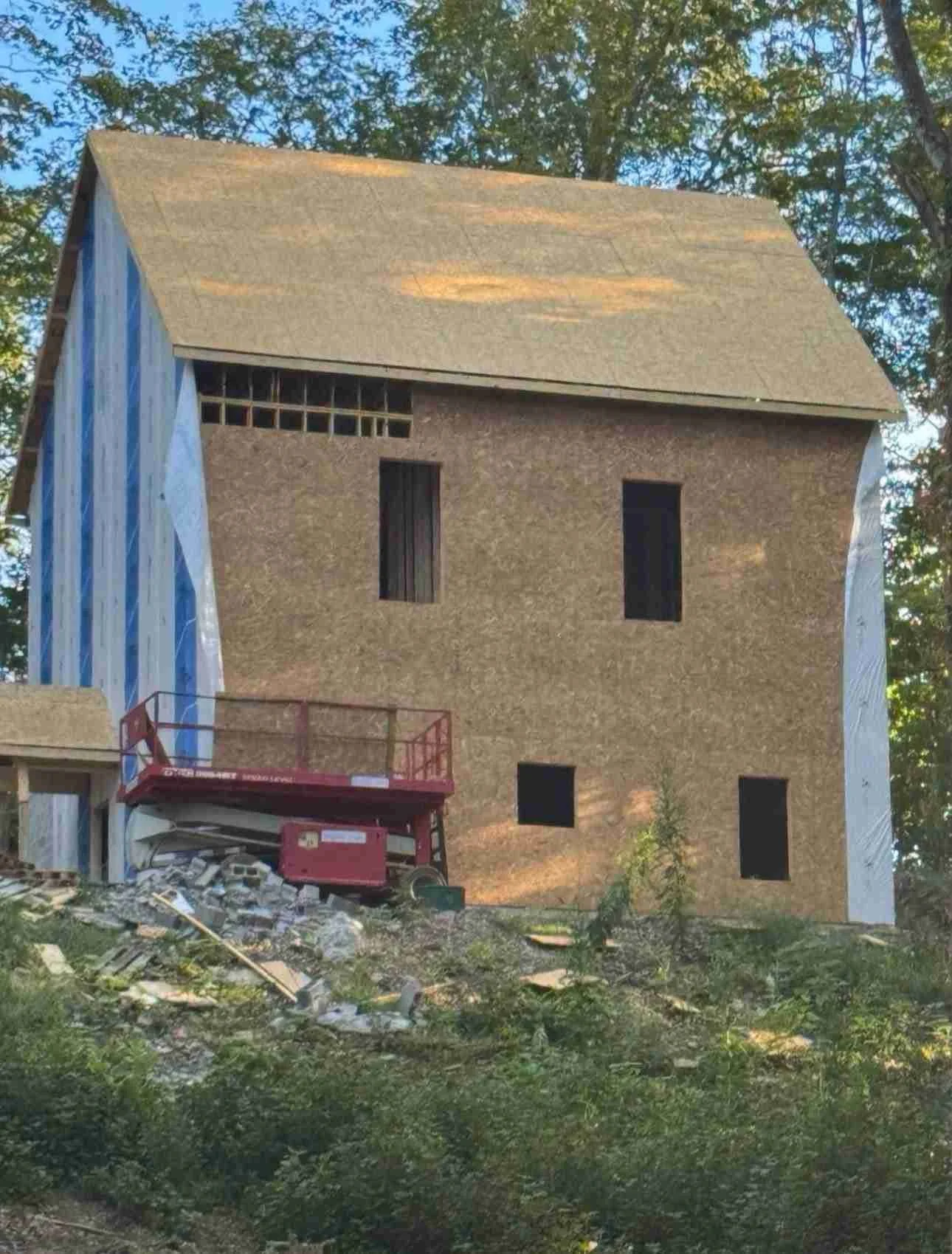 A house under construction with a partially completed wall and an unfinished exterior, surrounded by construction debris and a red lift, in a wooded area.