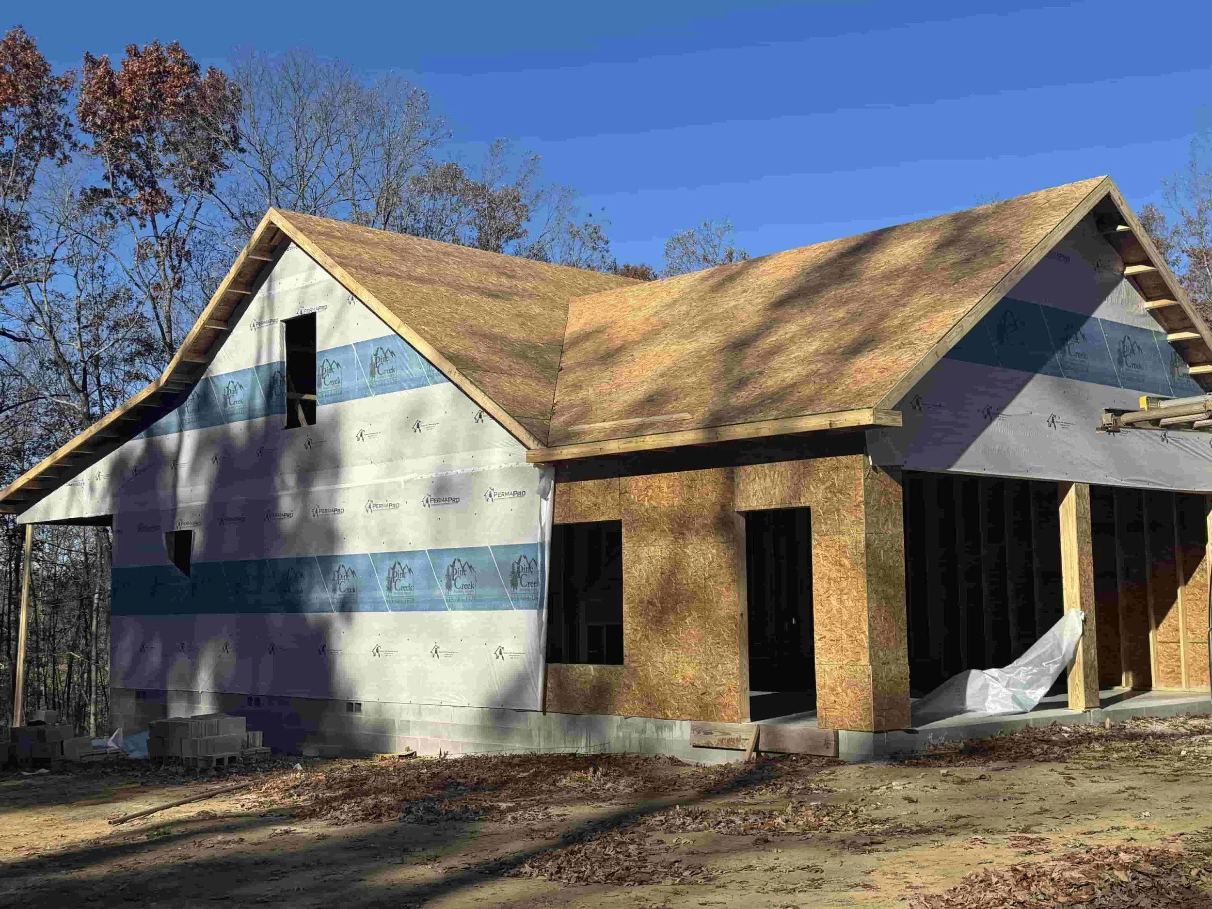 A house under construction with a partially completed roof and exterior walls, with trees in the background and a clear blue sky.