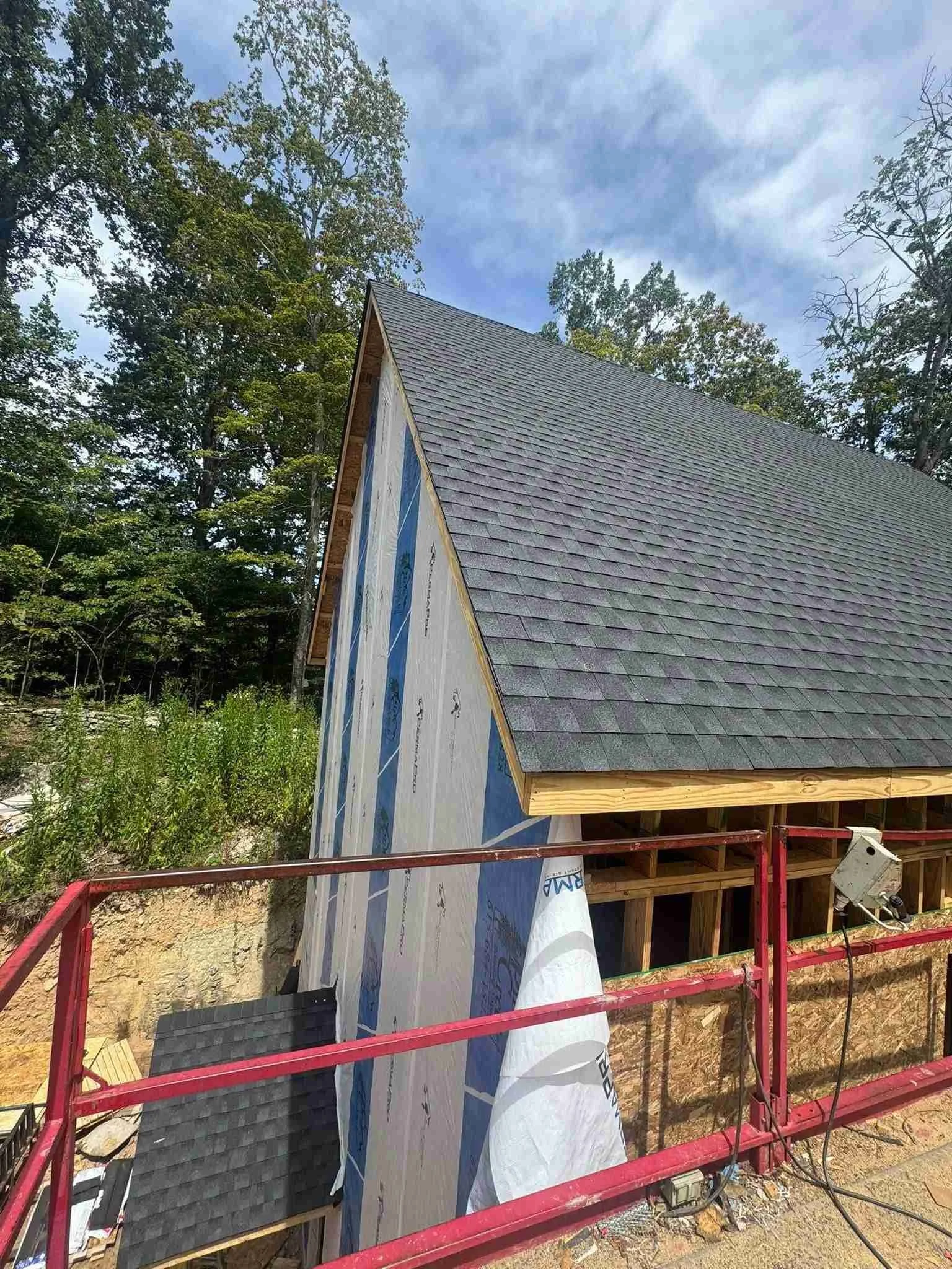 Construction site of a house with a steep roof, partially covered in dark shingles. The side wall has house wrapping material. There is a red safety railing in the foreground and green trees in the background.