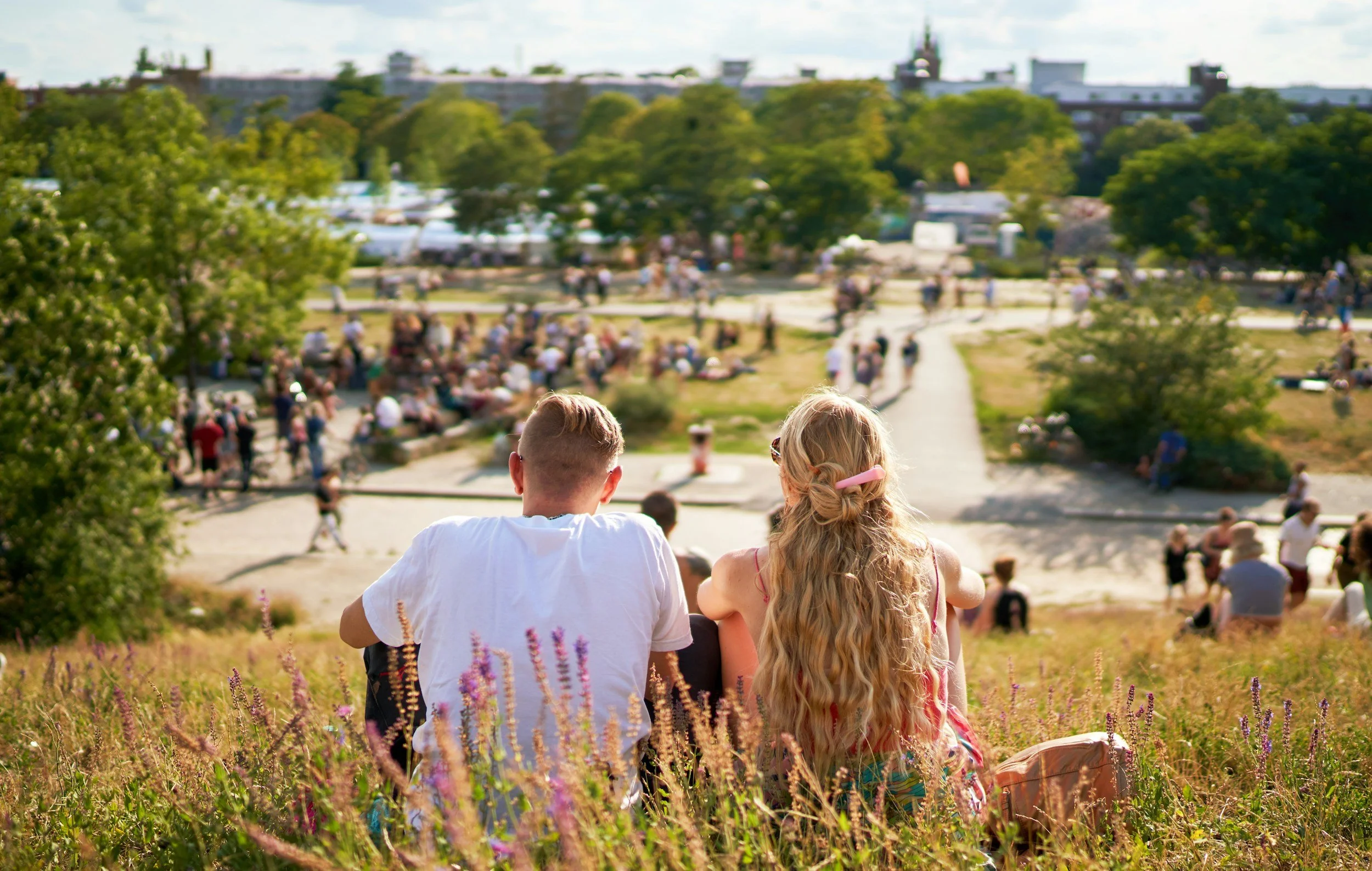 A young boy and girl sitting on a grassy hill overlooking a crowded park with many people relaxing and walking, trees, and buildings in the background on a sunny day.