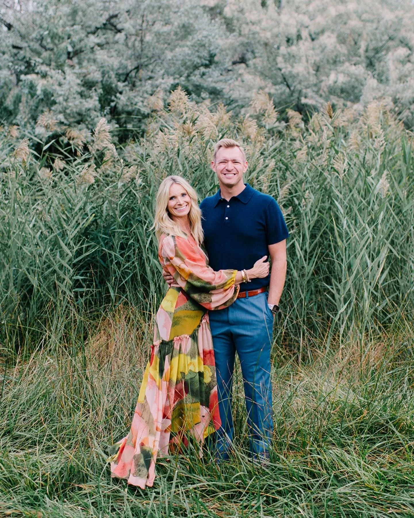 A smiling couple standing in a grassy field with tall green plants and trees in the background.