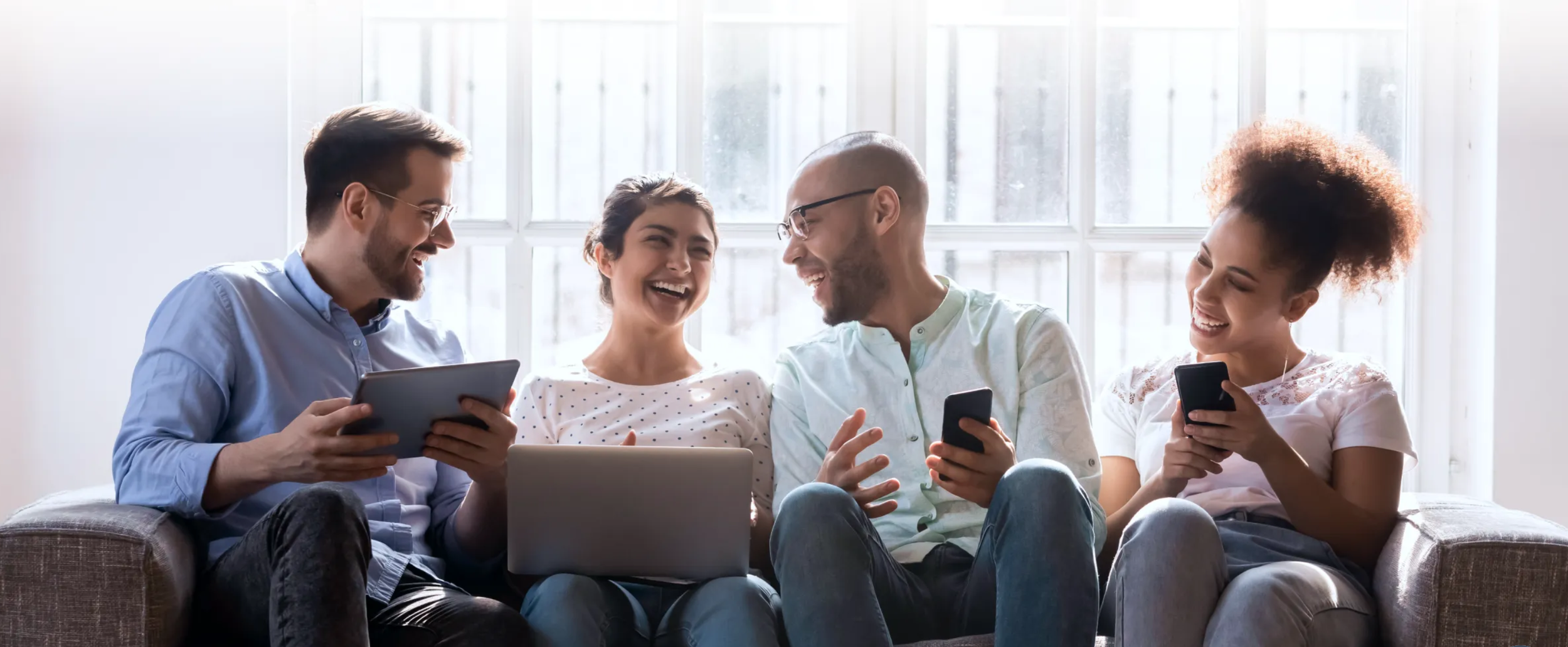 Group of four diverse young adults sitting on a couch, laughing and using electronic devices together in a bright room with large windows.