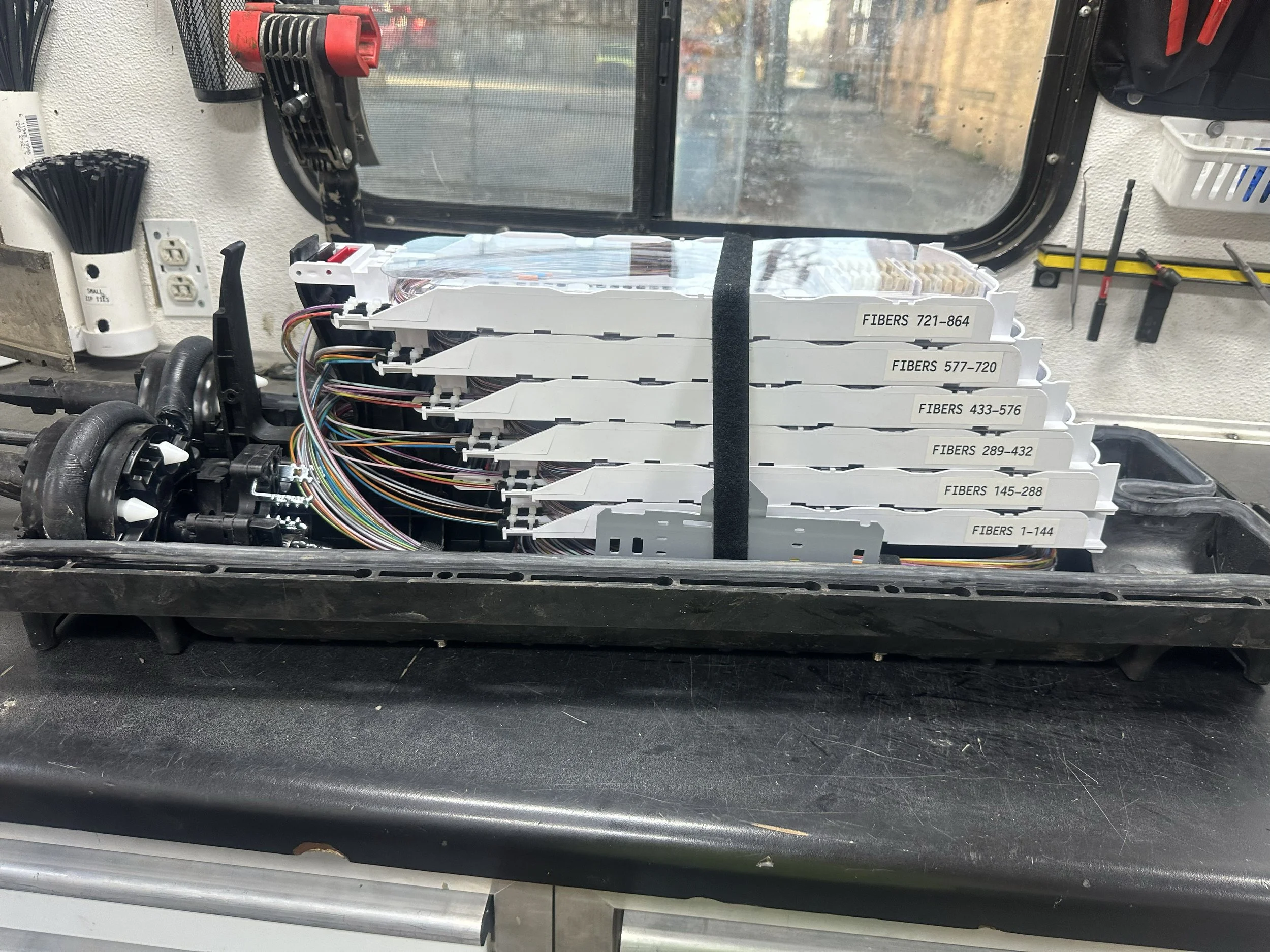 Open electronic device showing fiber optic modules labeled with fiber ranges and multi-colored cables connected inside a black plastic casing, on a workbench in a workshop.