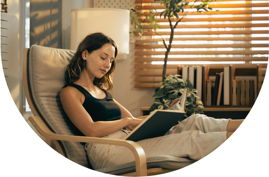 Mujer leyendo un libro en una silla en una habitación con cortinas de madera y una planta
