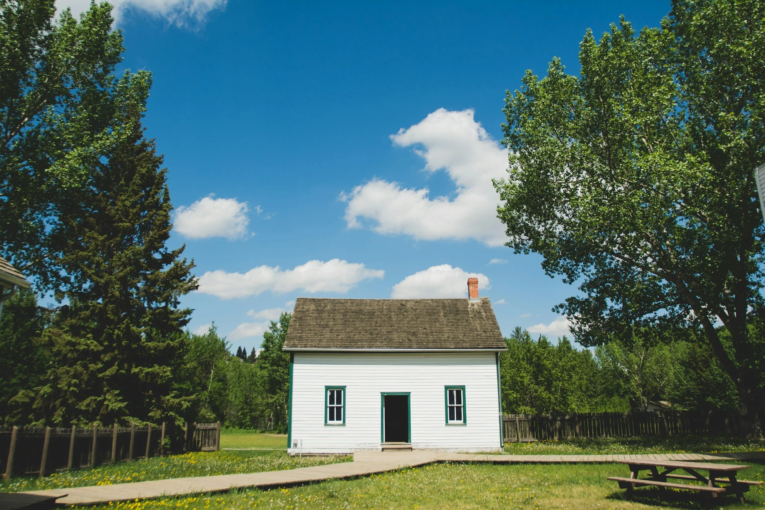Casa blanca de dos pisos en un jardín con árboles verdes y cielo azul con algunas nubes blancas.