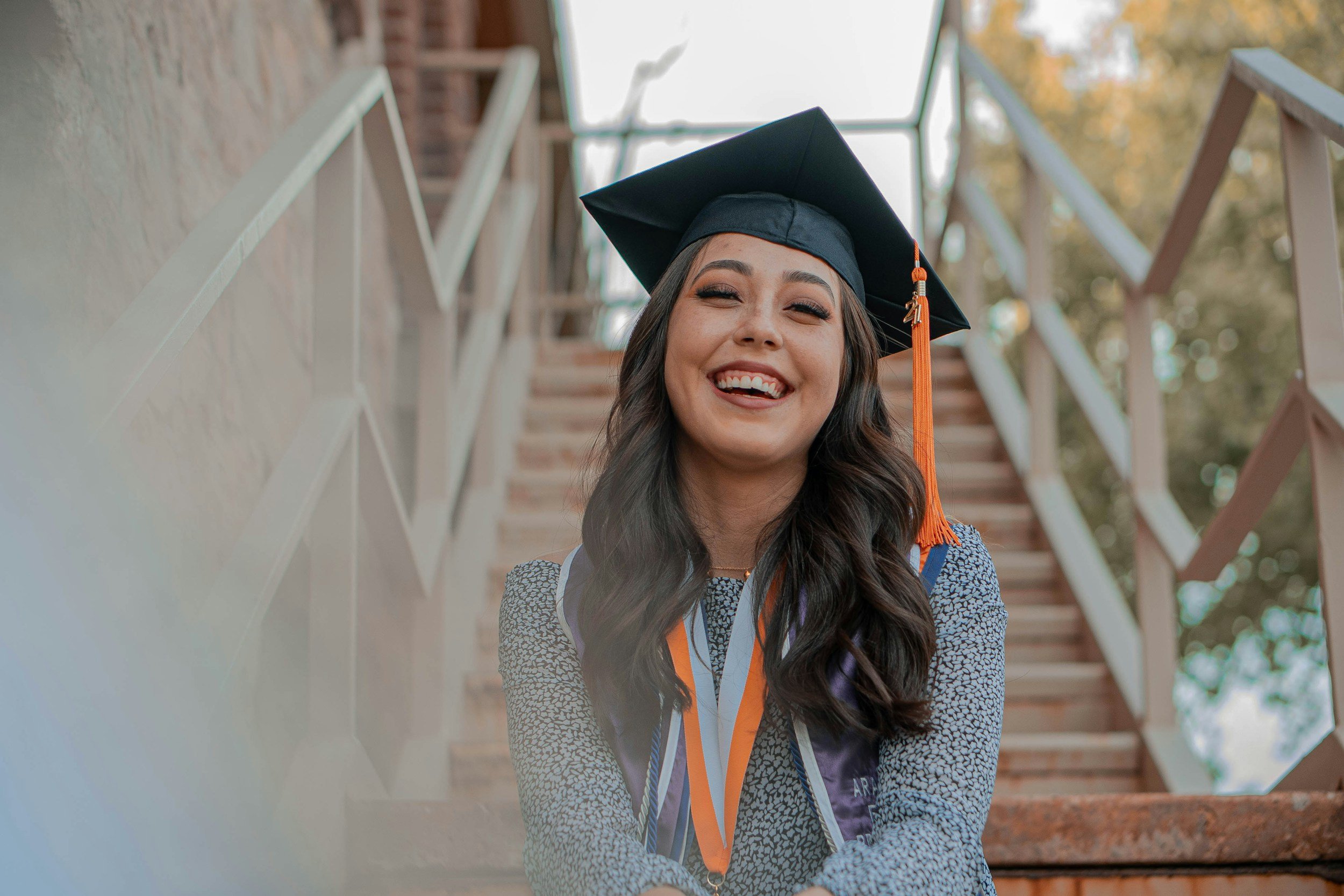 Joven graduada con toga y birrete sonriendo en un lugar al aire libre frente a una escalera de madera.