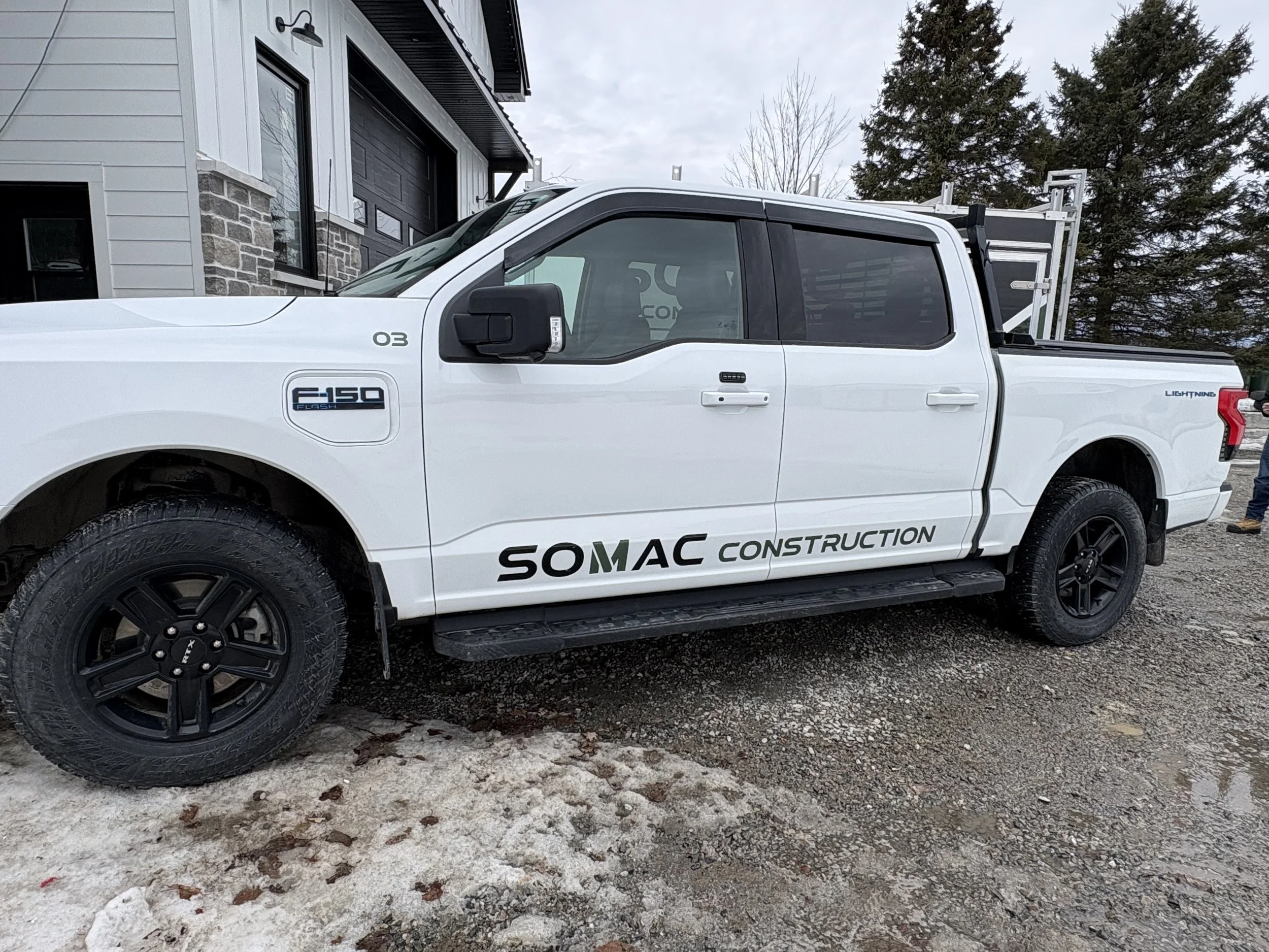 Camion blanc avec logo SOMAC Construction garé sur une surface de gravier et de neige, avec une maison moderne en arrière-plan.