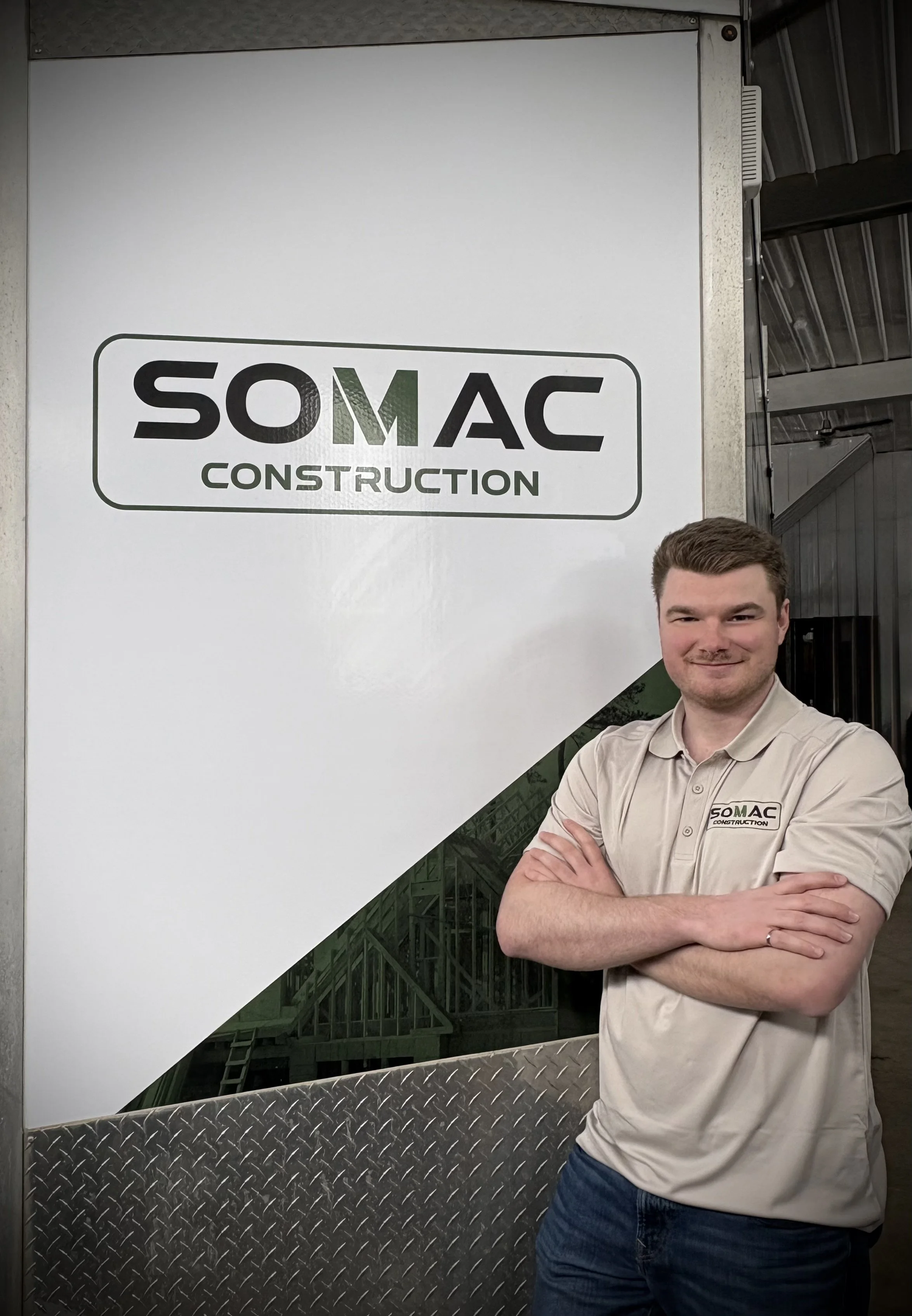 Un homme souriant en chemise beige portant un badge de la société SOMAC Construction, debout devant un panneau blanc avec le logo de SOMAC Construction, dans un environnement industriel.