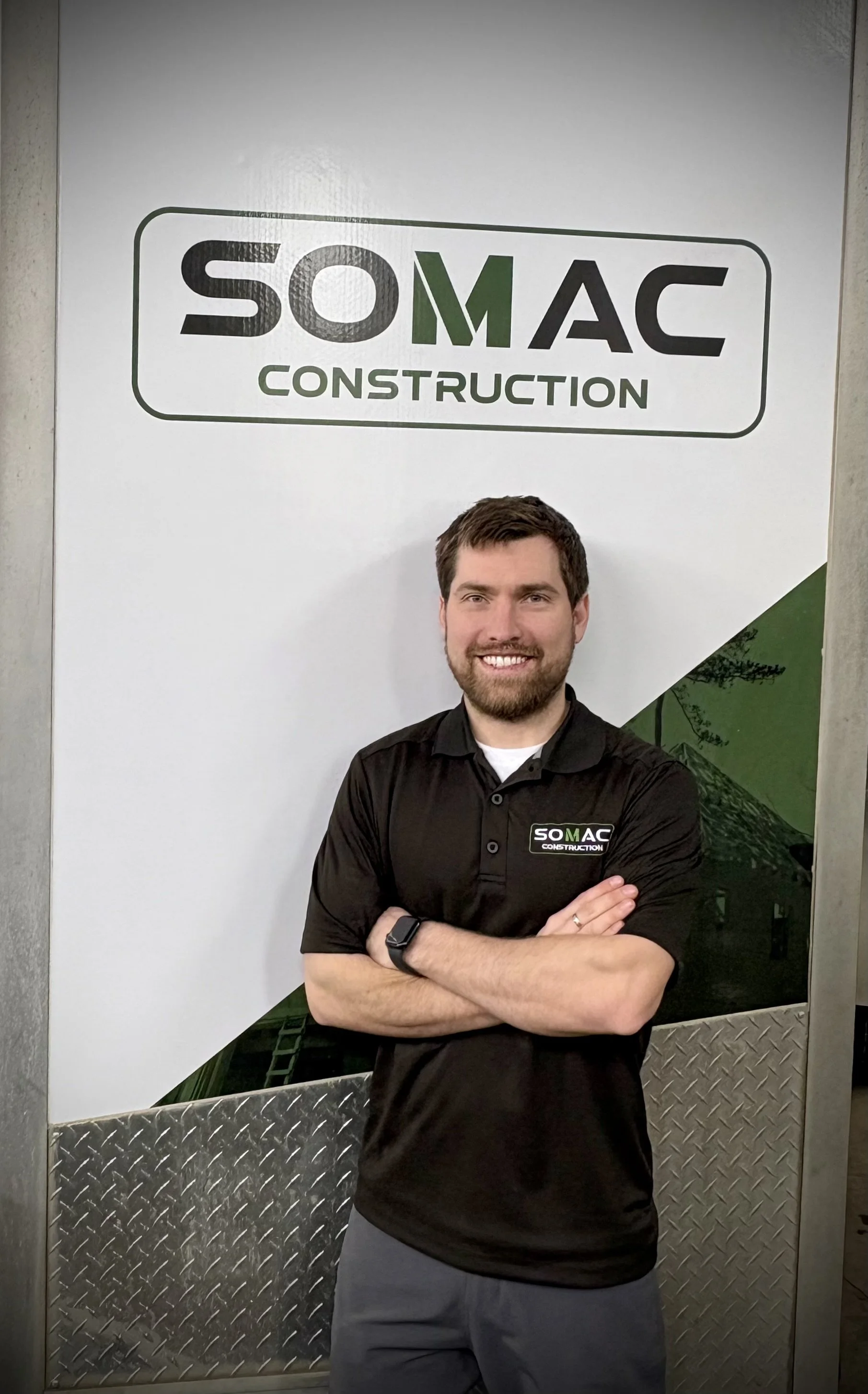 Un homme souriant avec un bras croisé portant un polo noir avec le logo SOMAC CONSTRUCTION devant un panneau blanc avec le logo SOMAC CONSTRUCTION.