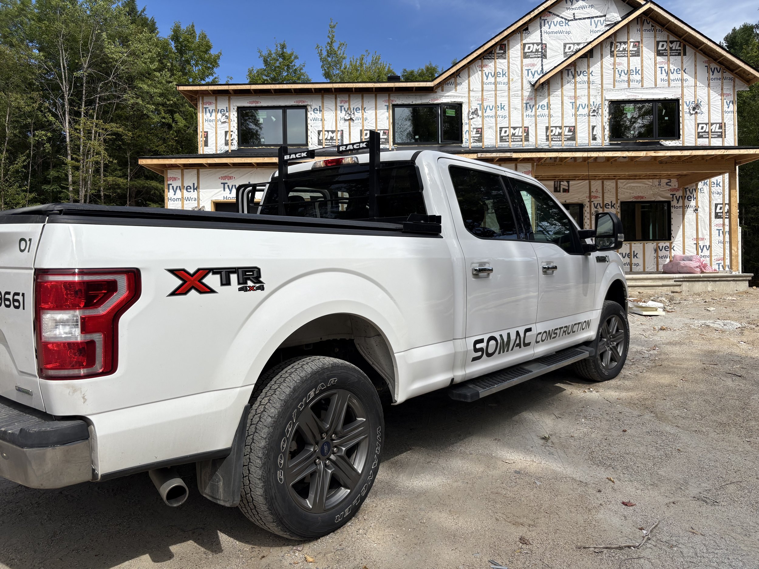 Camion blanc de construction avec le logo SOMAC Construction et XTR 4X4 garé devant une maison en construction, le bâtiment est encore en structure avec des murs en osb et des fenêtres installées, entouré d'arbres et un ciel bleu.
