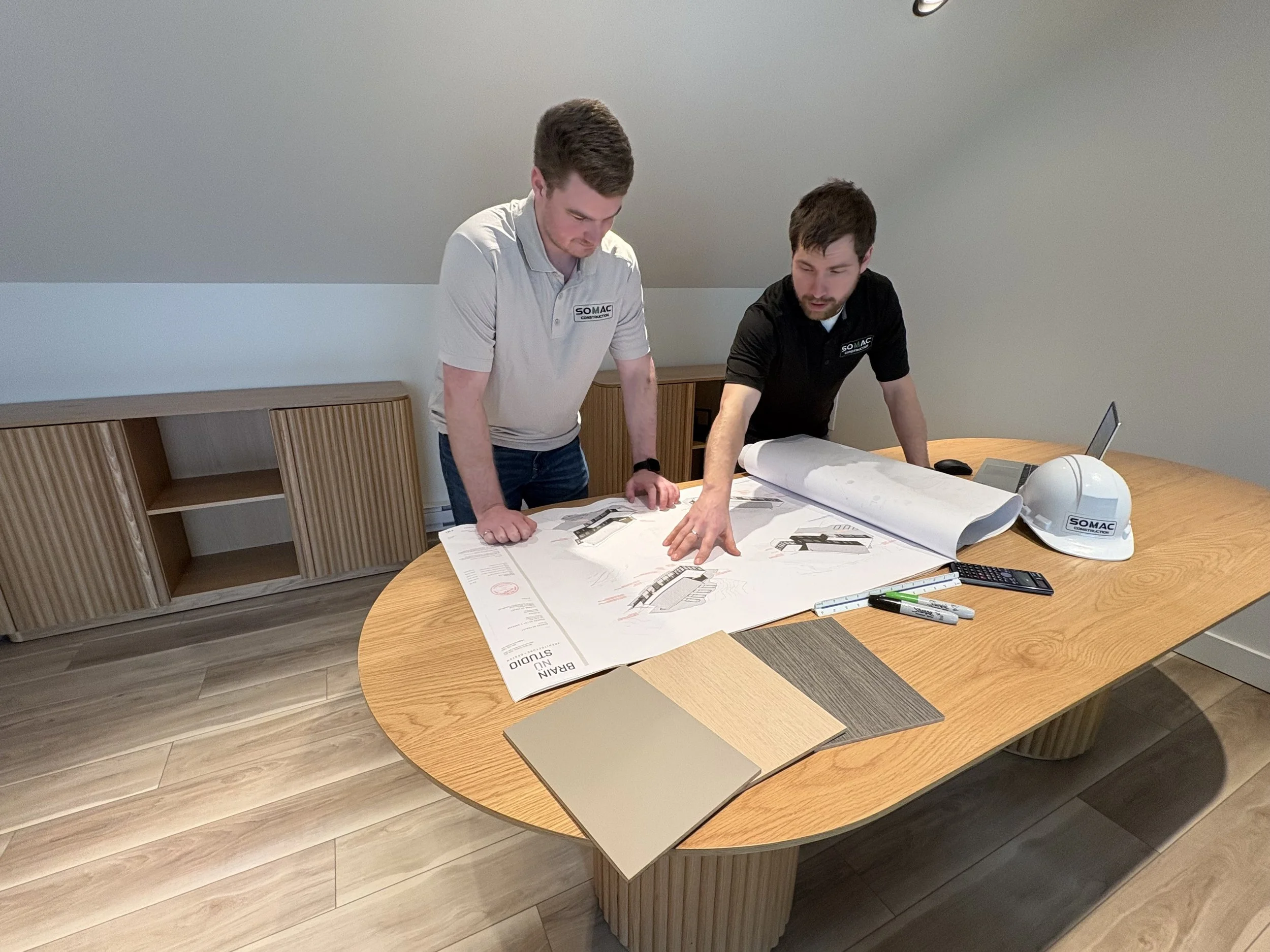Deux hommes en polo travaillant sur une grande feuille de plans architecturaux sur une table en bois, avec des échantillons de matériaux et des outils de dessin.