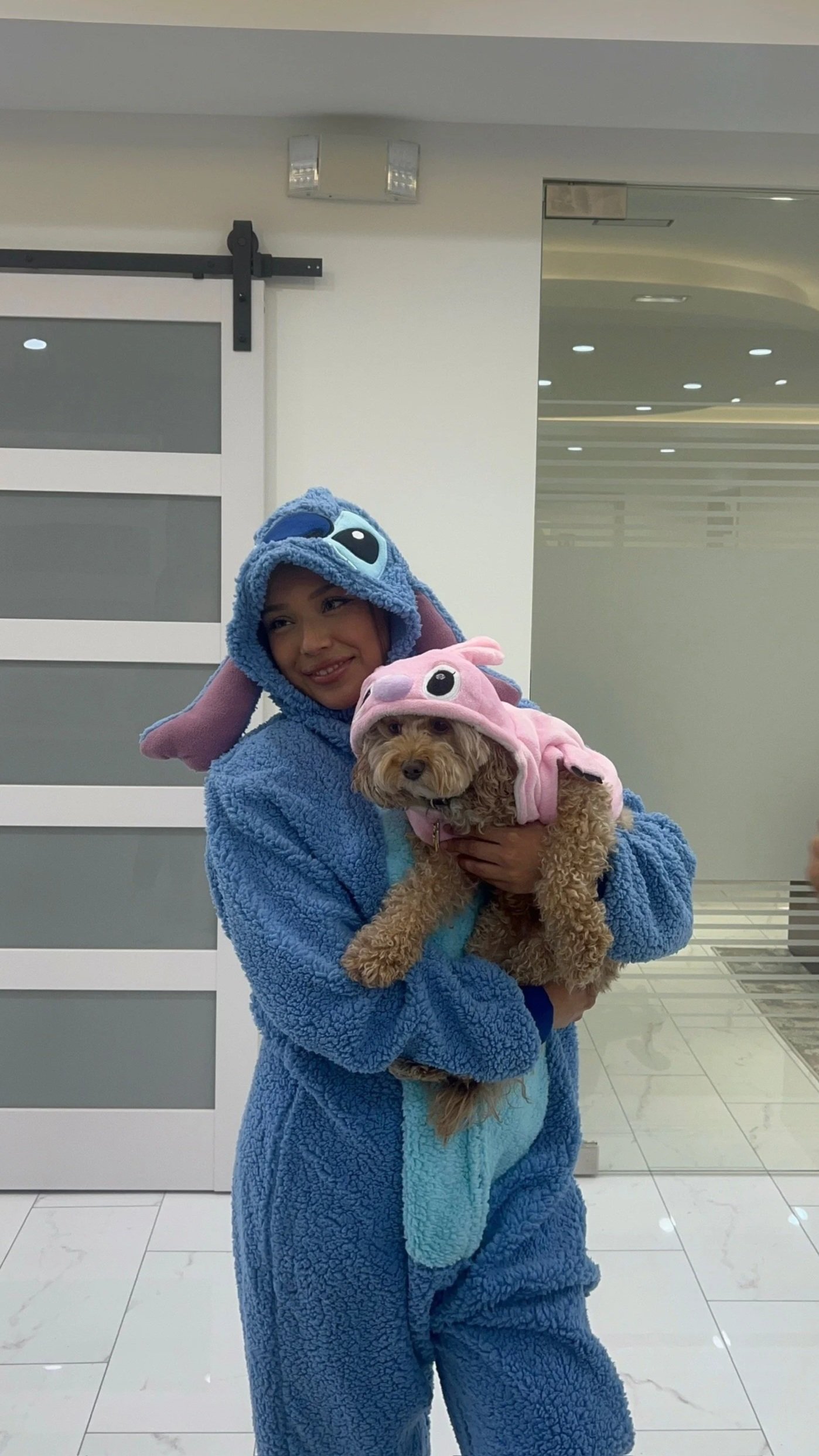 A woman dressed in a blue Stitch costume from the movie Lilo & Stitch, holding a small dog dressed in a pink bunny costume. They are indoors near a sliding door.