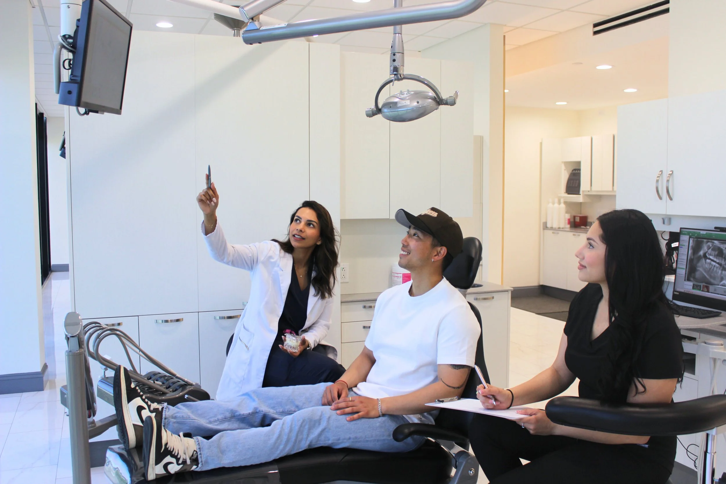 A young man lying on a dental chair with a woman taking a selfie and two women watching, in a dental clinic.