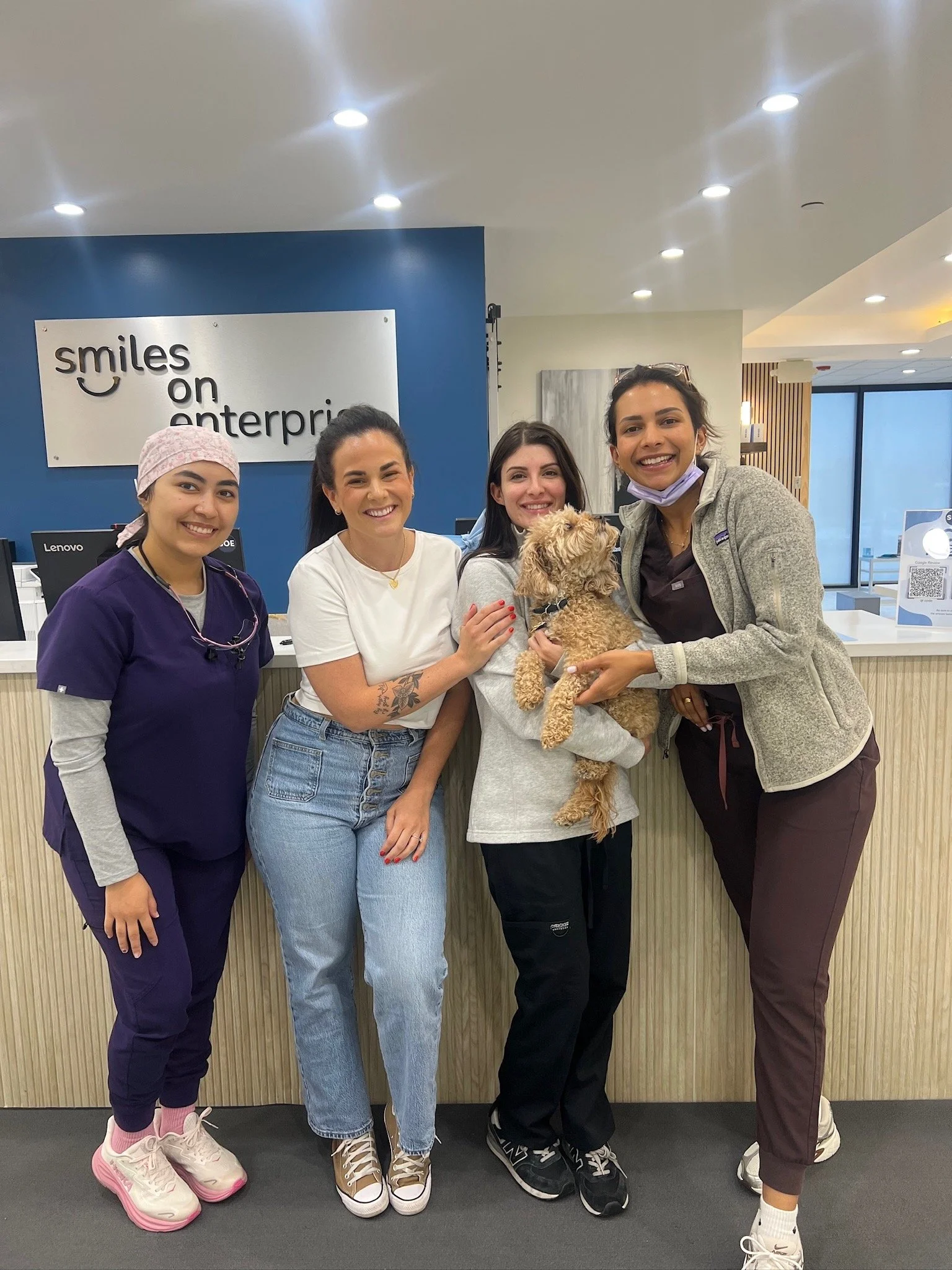 Four women standing at a reception desk, with one woman holding a small dog. The background has a sign that reads 'smiles on enterprise'.