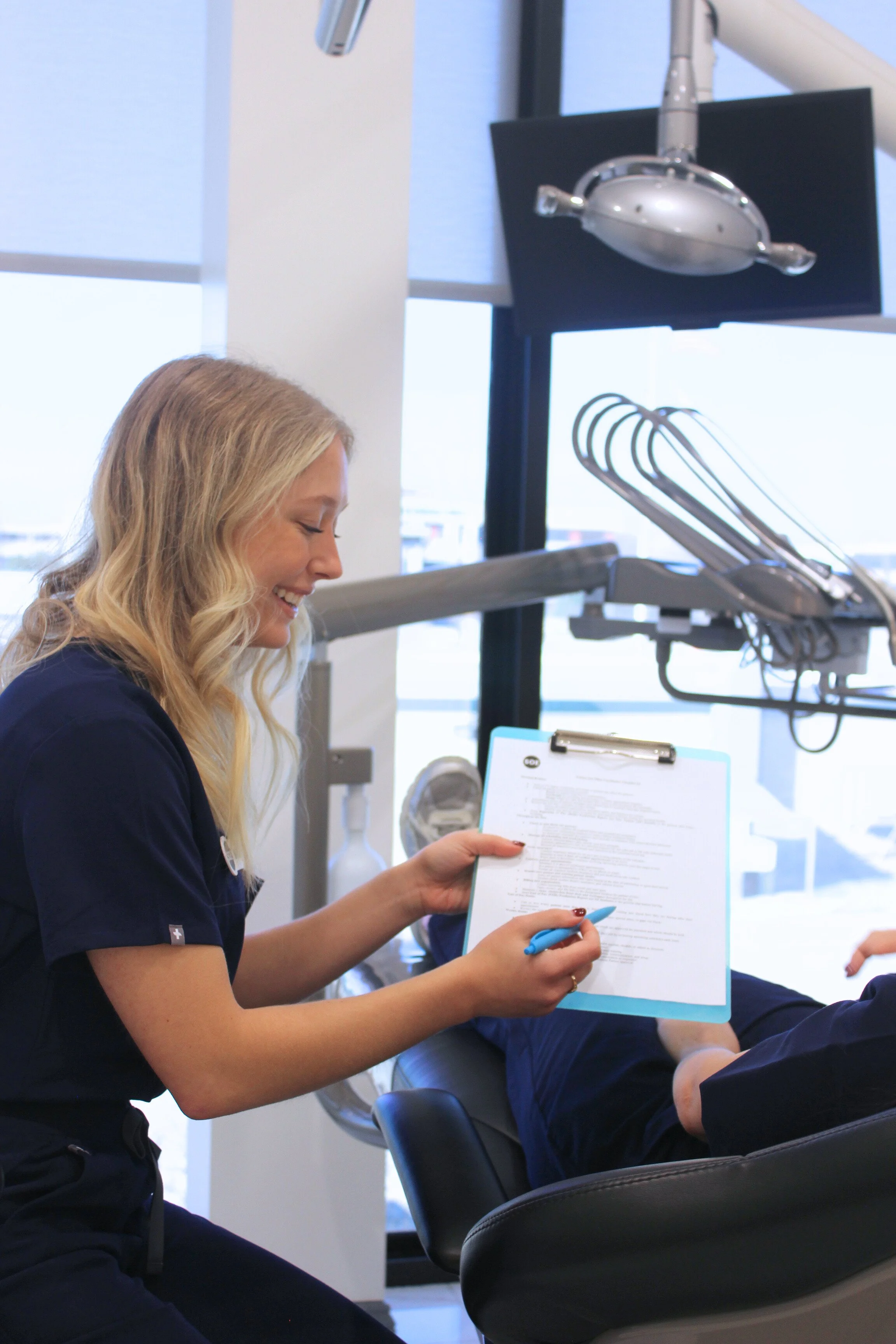 A female dentist smiling and holding a clipboard with paperwork while examining a patient reclining in a dental chair in a modern dental clinic.