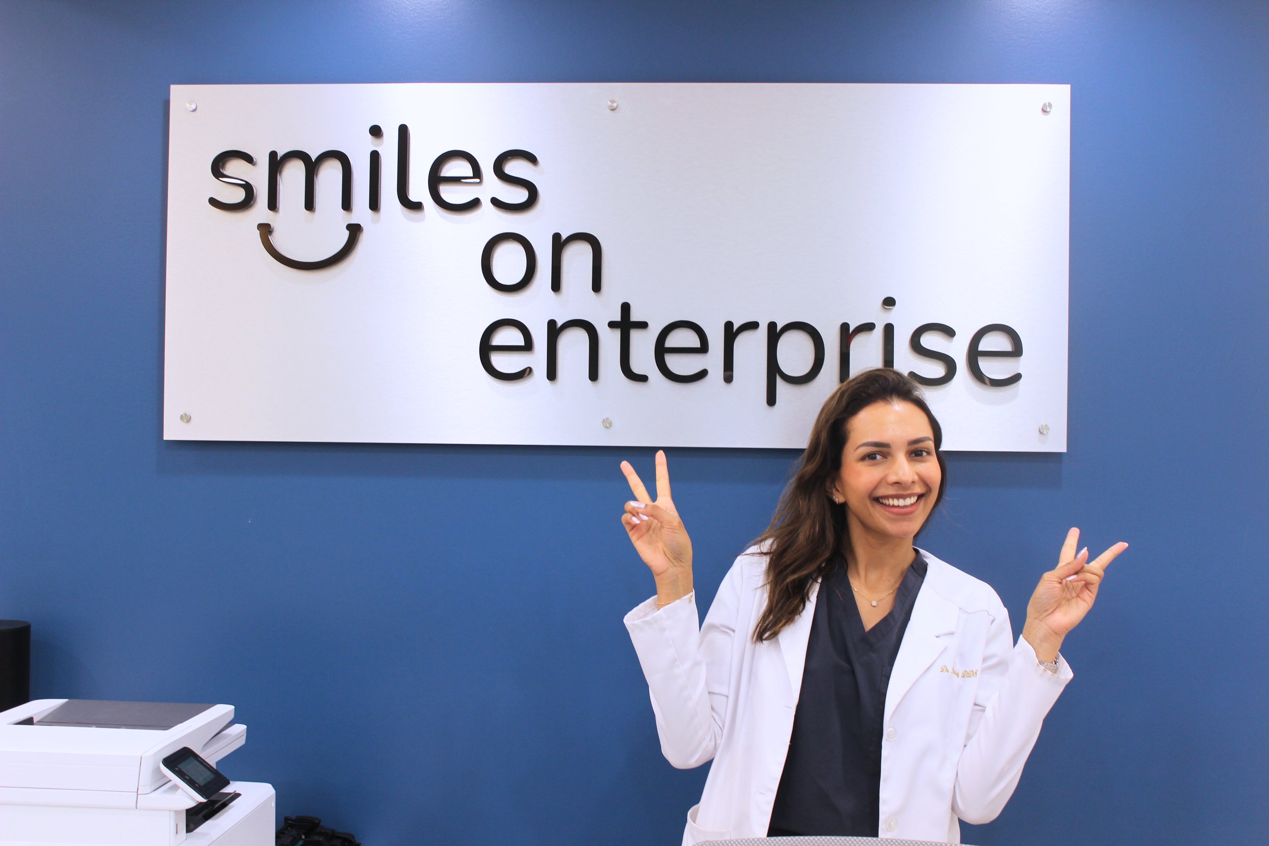A smiling woman in a medical coat making peace signs in front of a large sign that reads 'smiles on enterprise' on a blue wall.