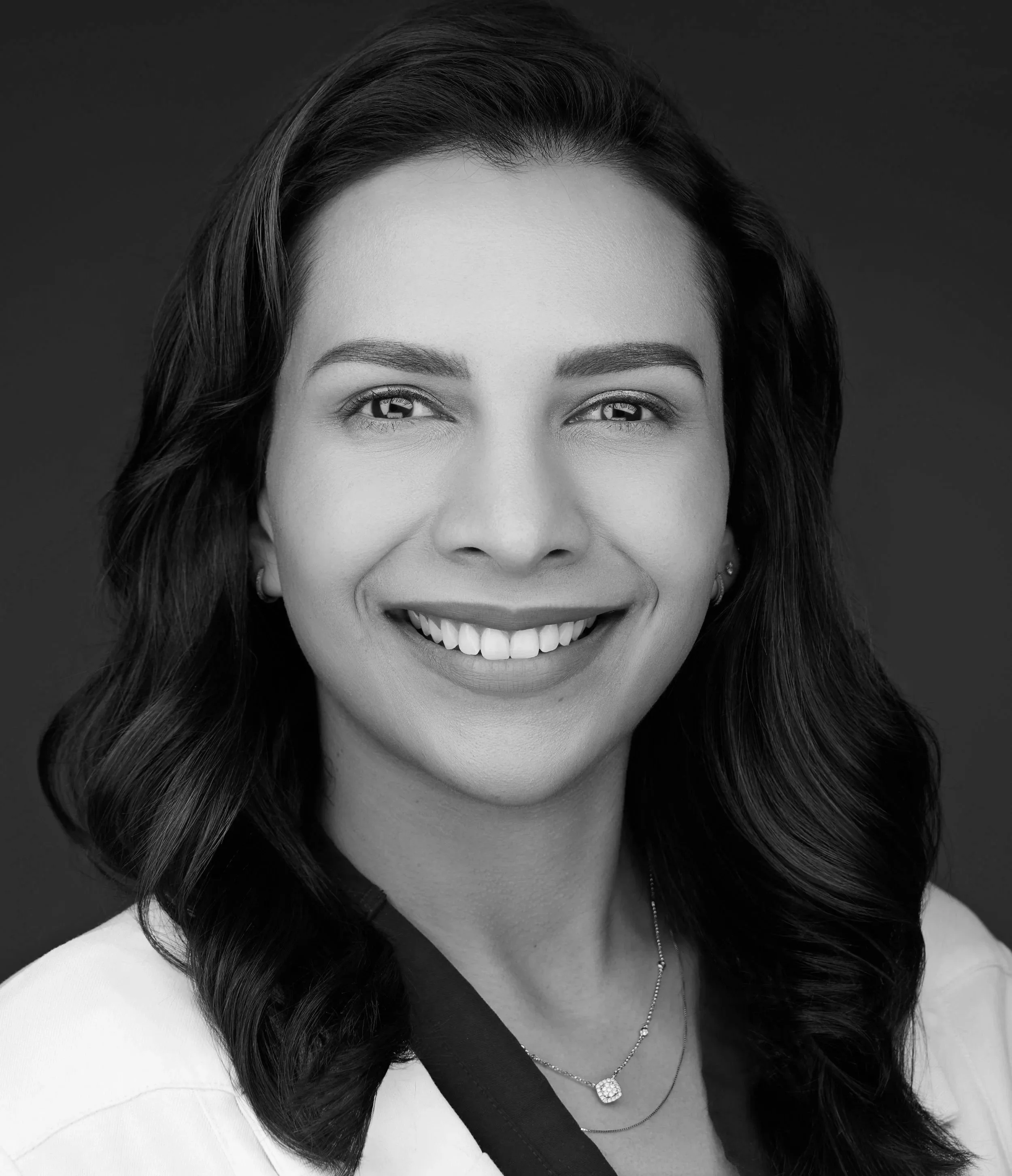 Black and white portrait of a smiling woman with wavy hair, wearing earrings, a necklace, and a light-colored top with a dark collar.