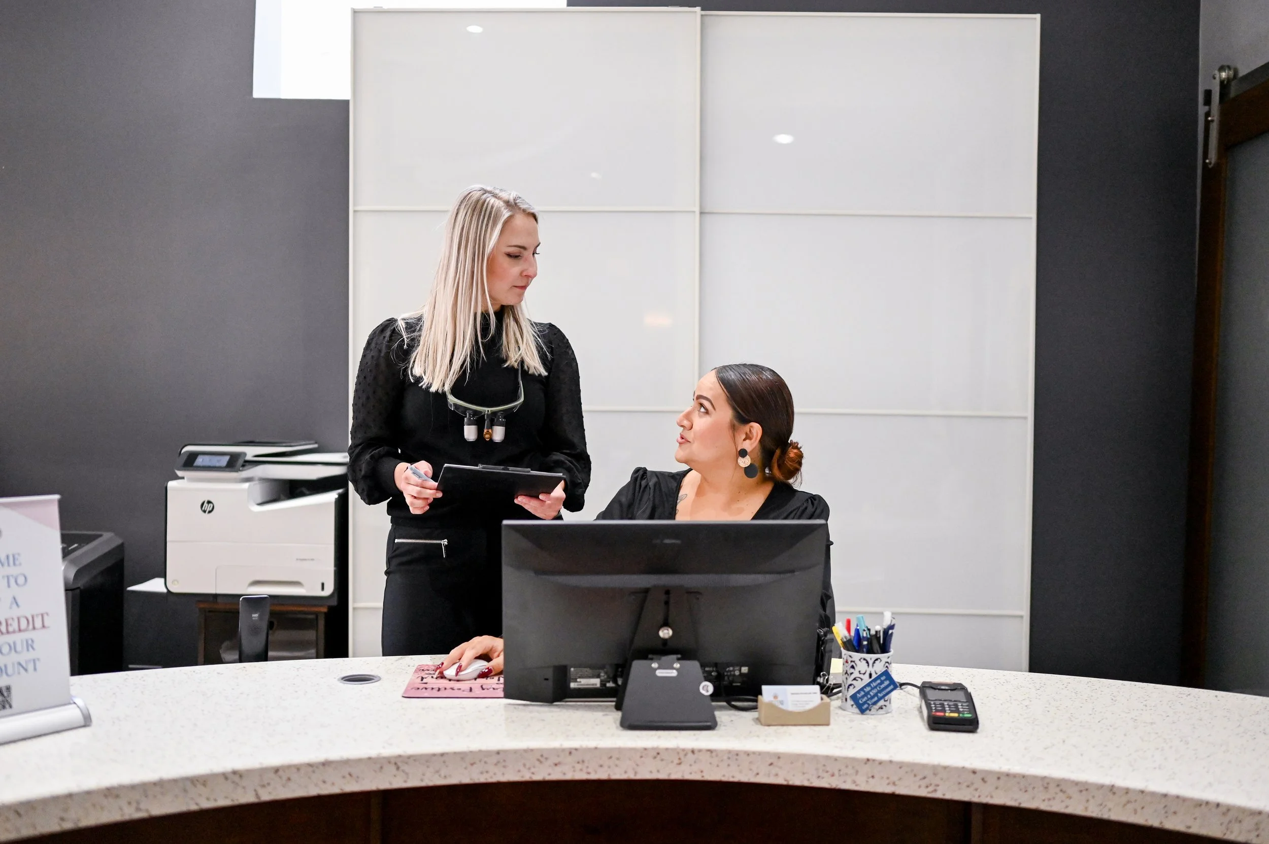 A woman standing and holding a clipboard talking to a woman sitting at a desk in an office.