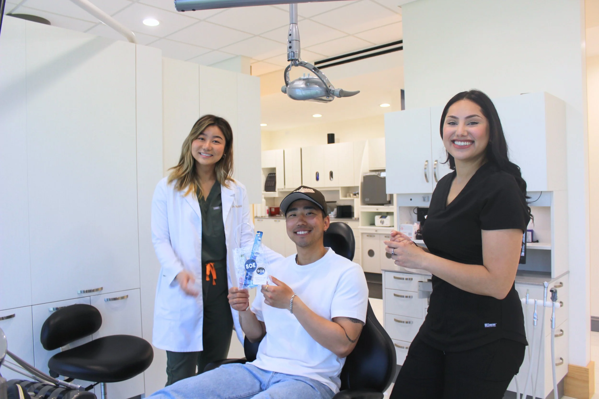 A man in a dental chair holding a small gift bag, flanked by two women in medical scrubs, inside a dental office.
