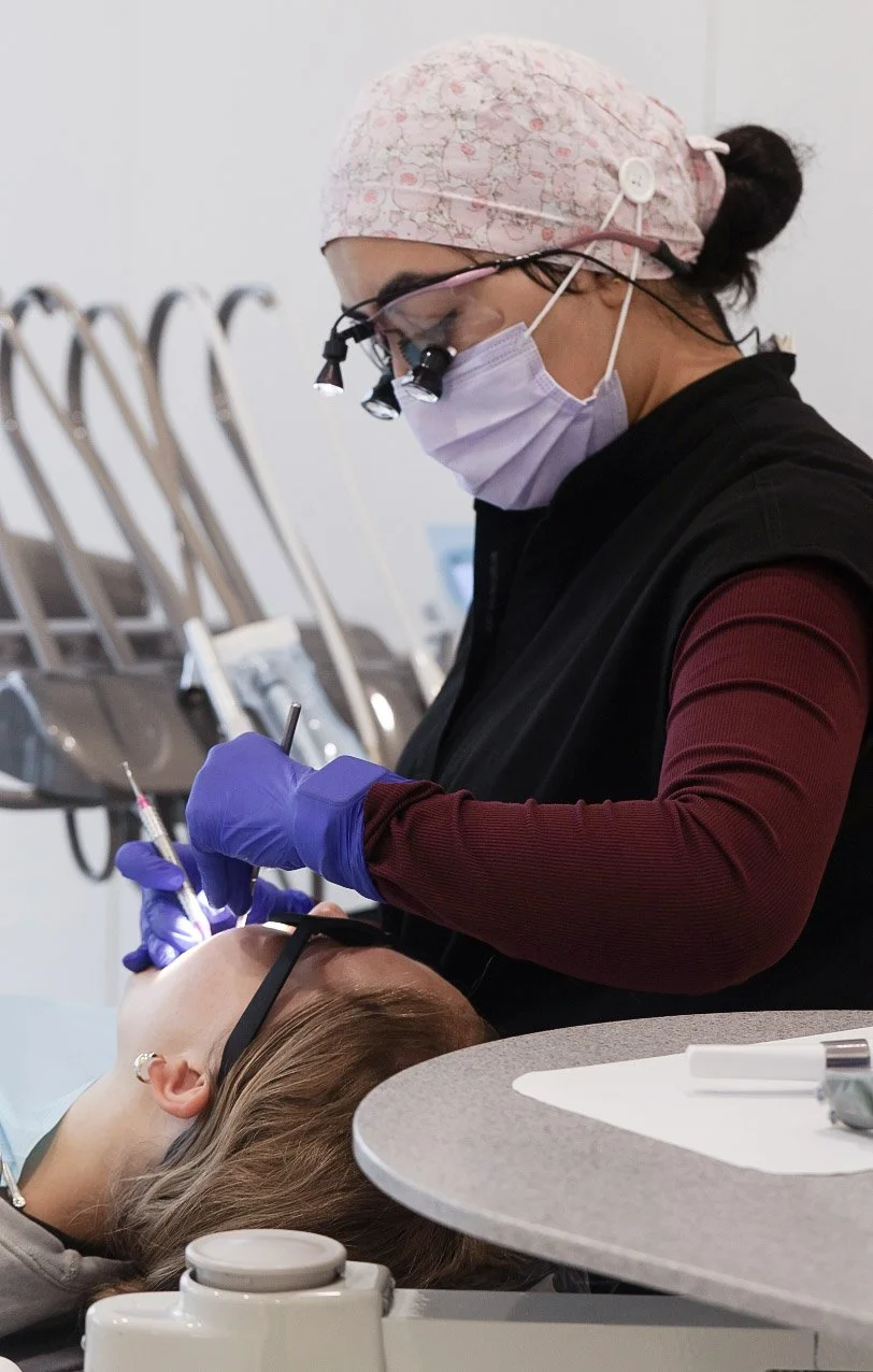 A dentist wearing magnifying glasses, a surgical mask, and gloves, treats a patient lying down in a dental chair.
