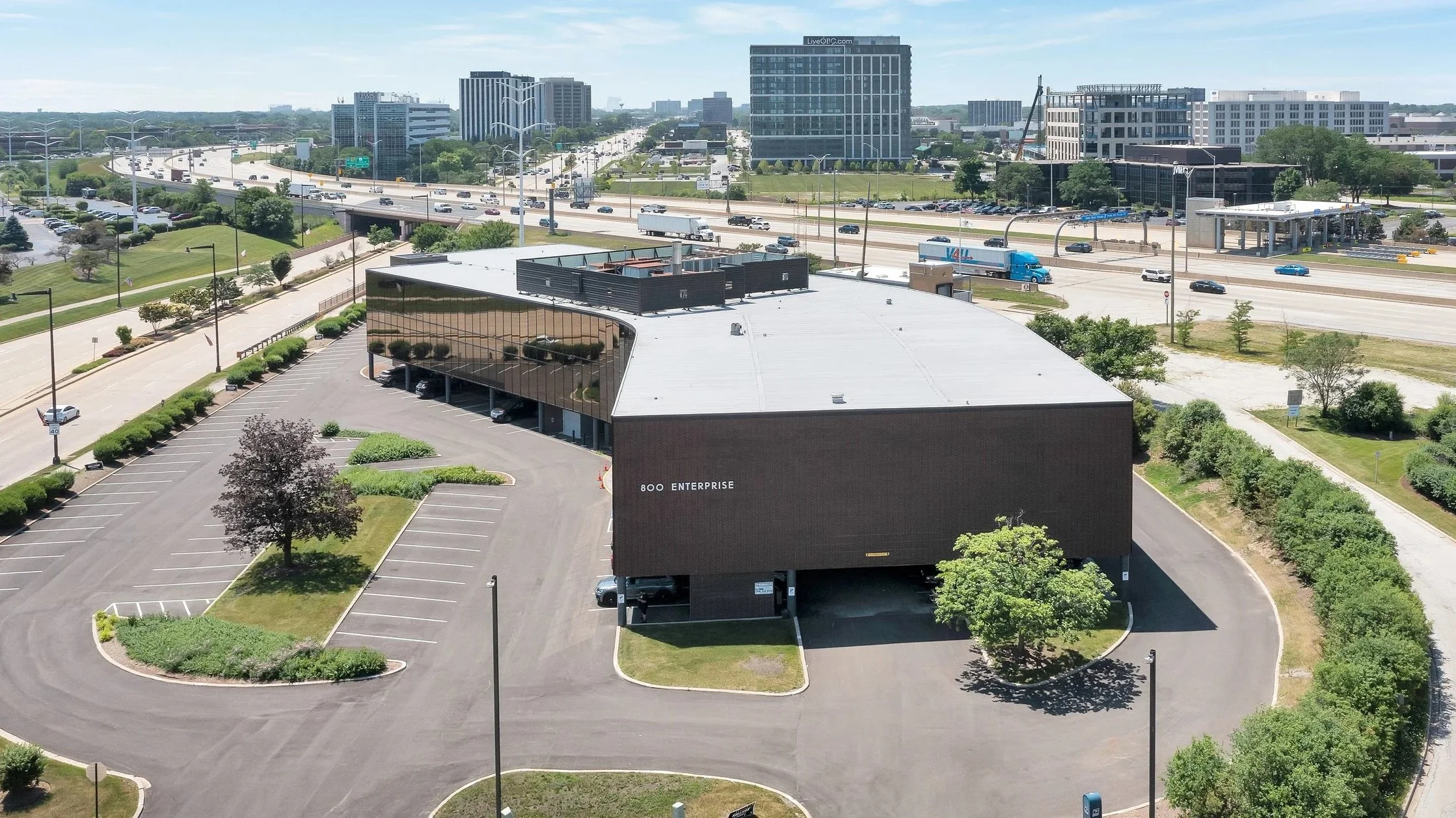 Aerial view of a commercial building with a large parking lot, surrounded by trees, with highways and city buildings in the background.