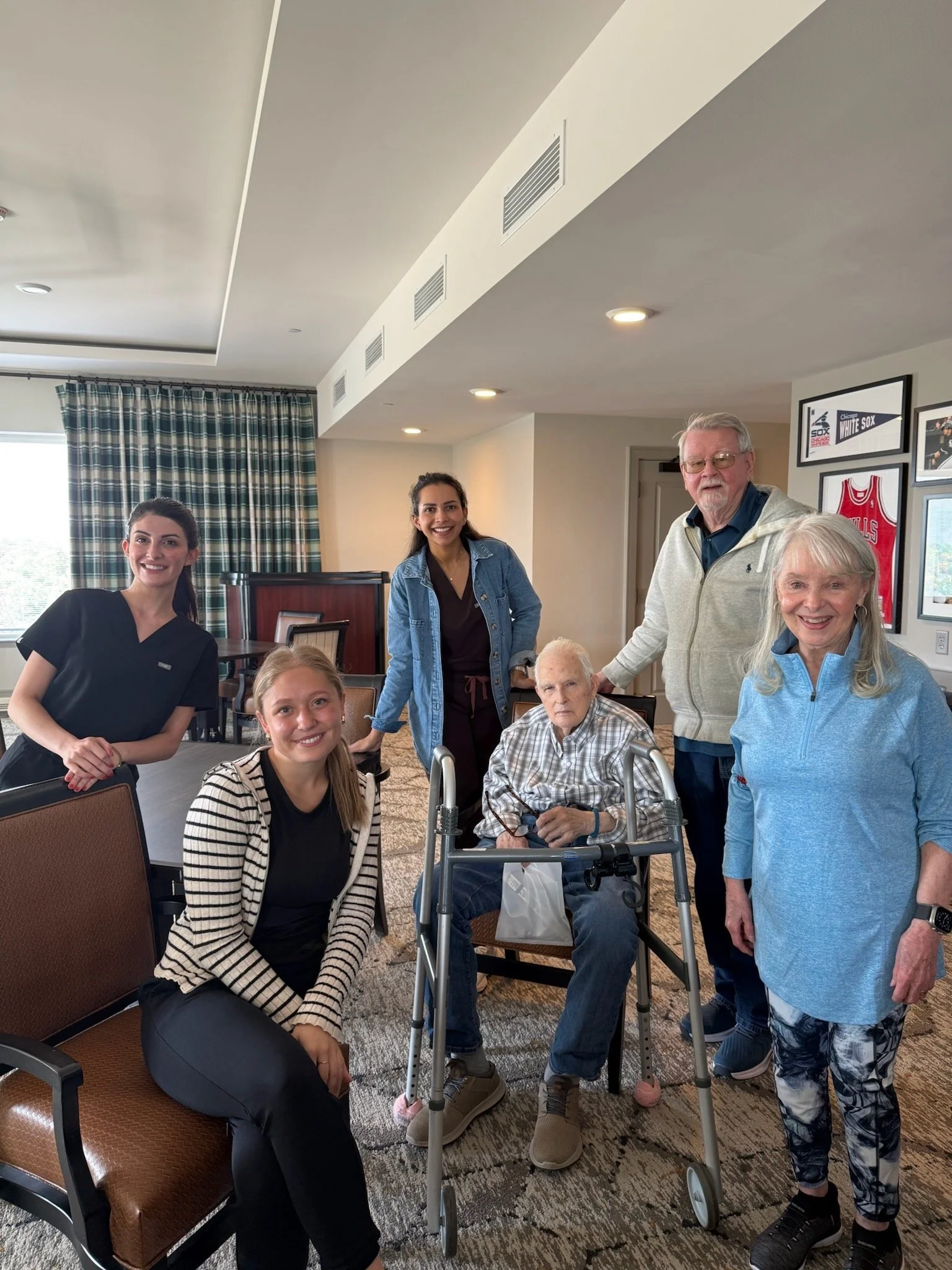 Group of six people, including an elderly man in a wheelchair, smiling in a room with sports memorabilia on the wall.