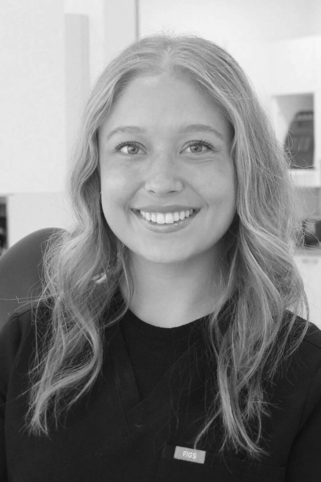 Black and white photo of a smiling woman with wavy hair wearing a uniform with a Figs logo, seated in an office environment.