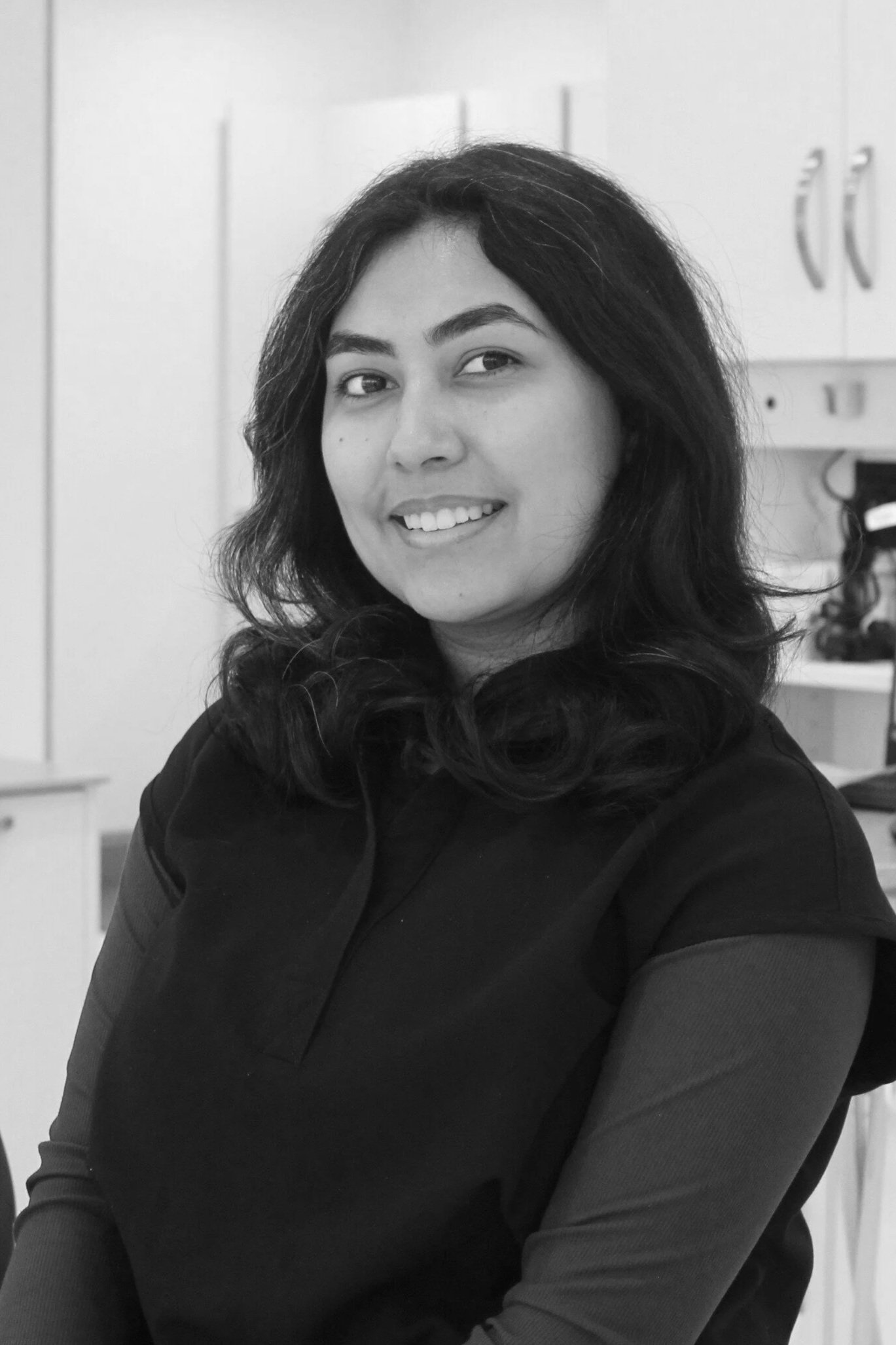 A woman with long, dark, wavy hair smiling and looking to the side, sitting in a room with cabinets and some equipment in the background.