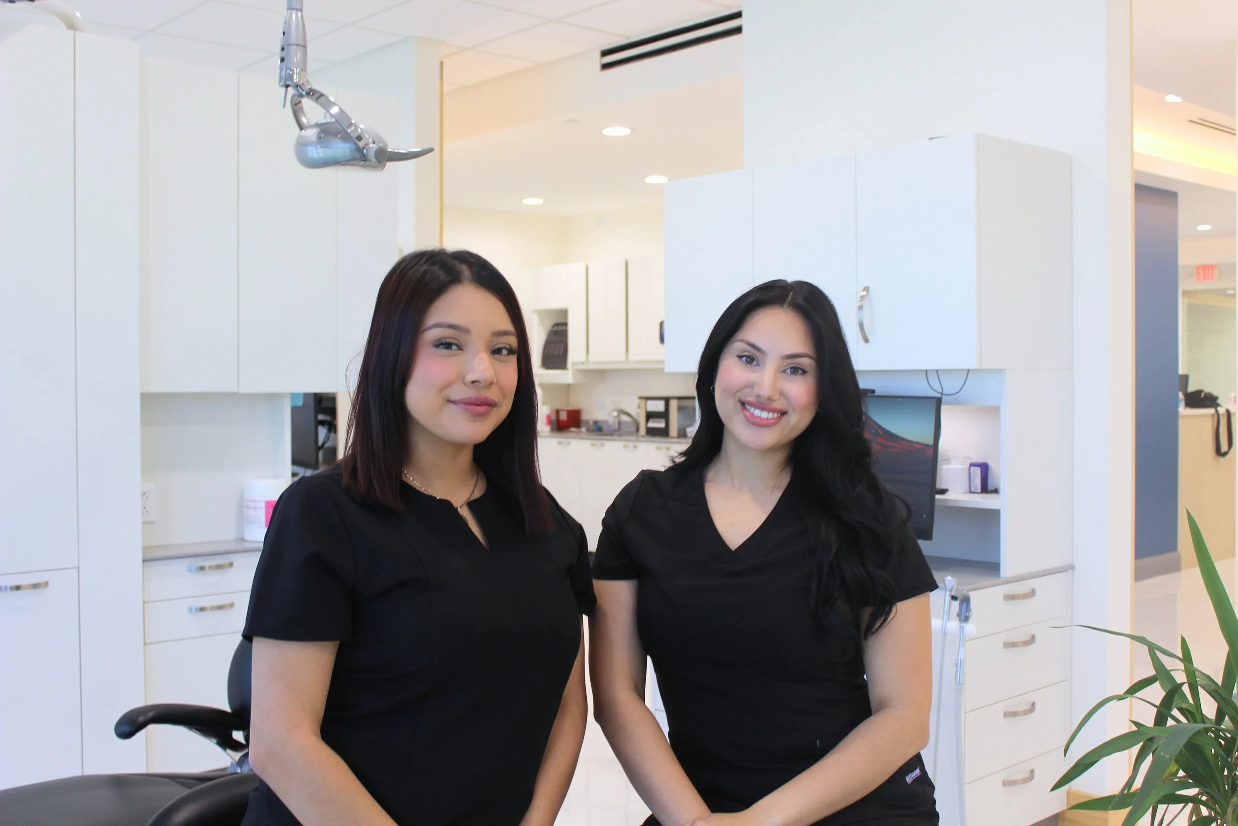 Two smiling women in black scrubs stand in a medical office with white cabinets and a computer in the background.