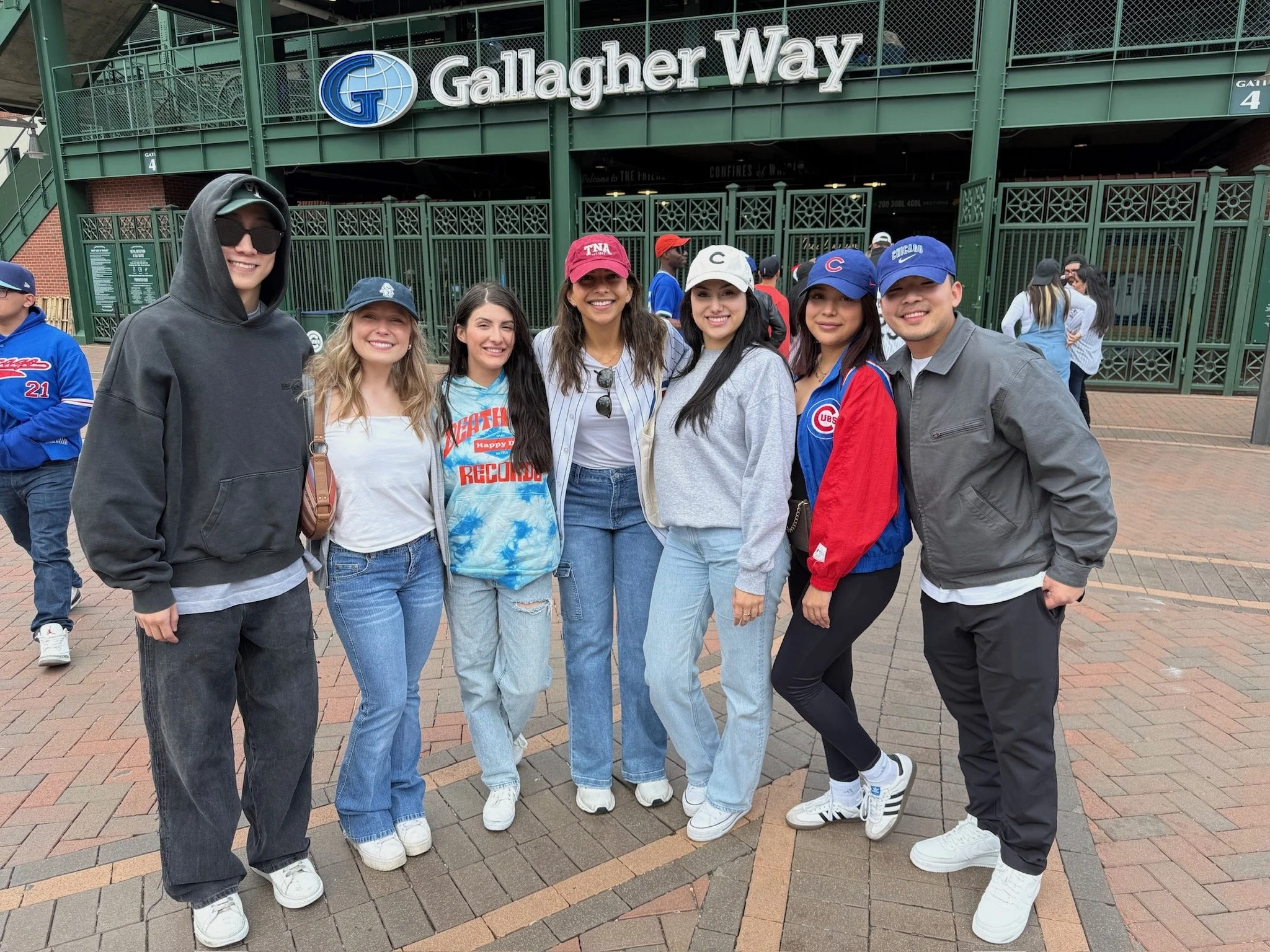 Group of seven young people posing in front of Gallagher Way entrance at Wrigley Field, Chicago, wearing casual sportswear and Chicago Cubs apparel.