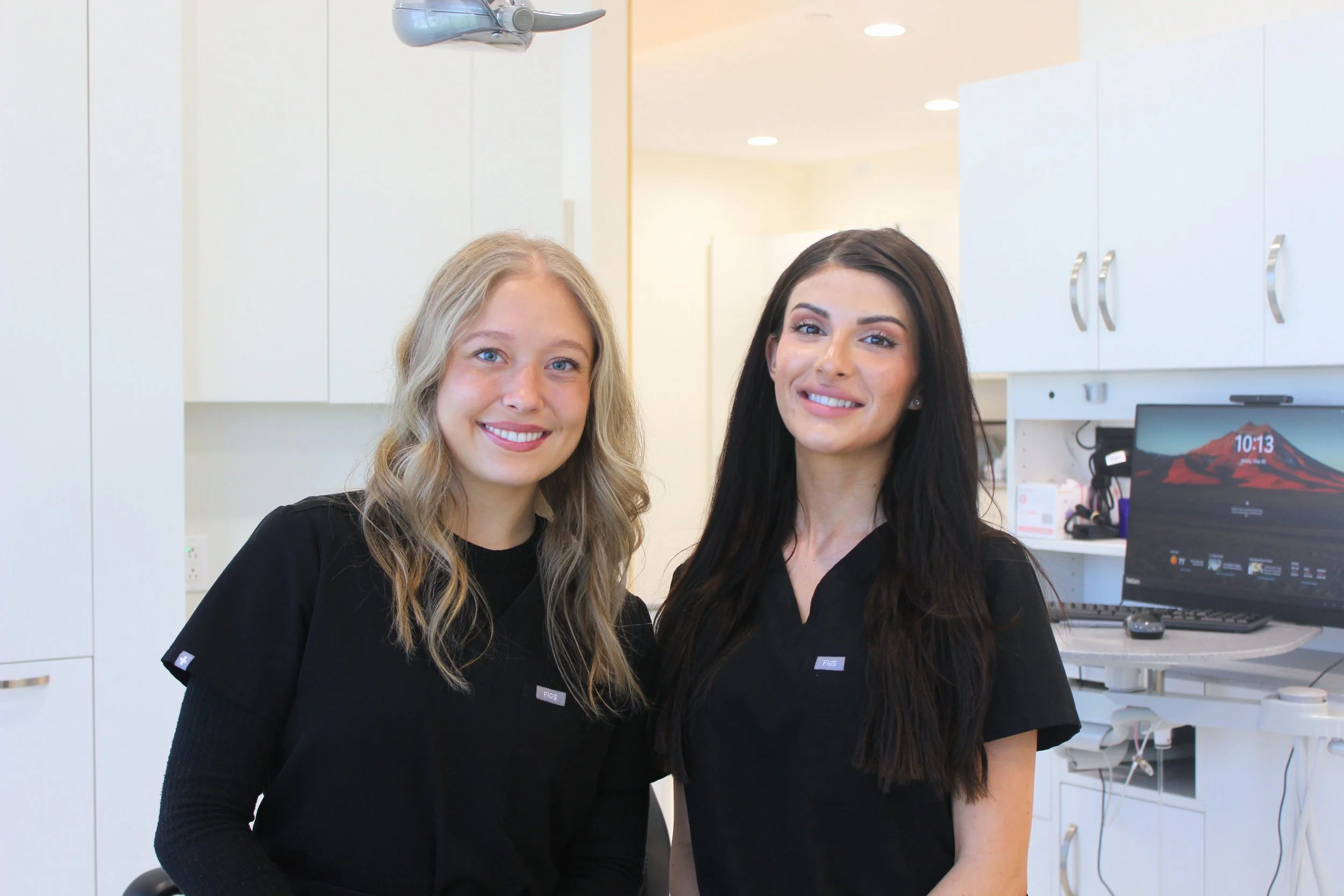 Two smiling dental professionals standing in a dental clinic, wearing black scrubs, with white cabinets and a computer monitor in the background.