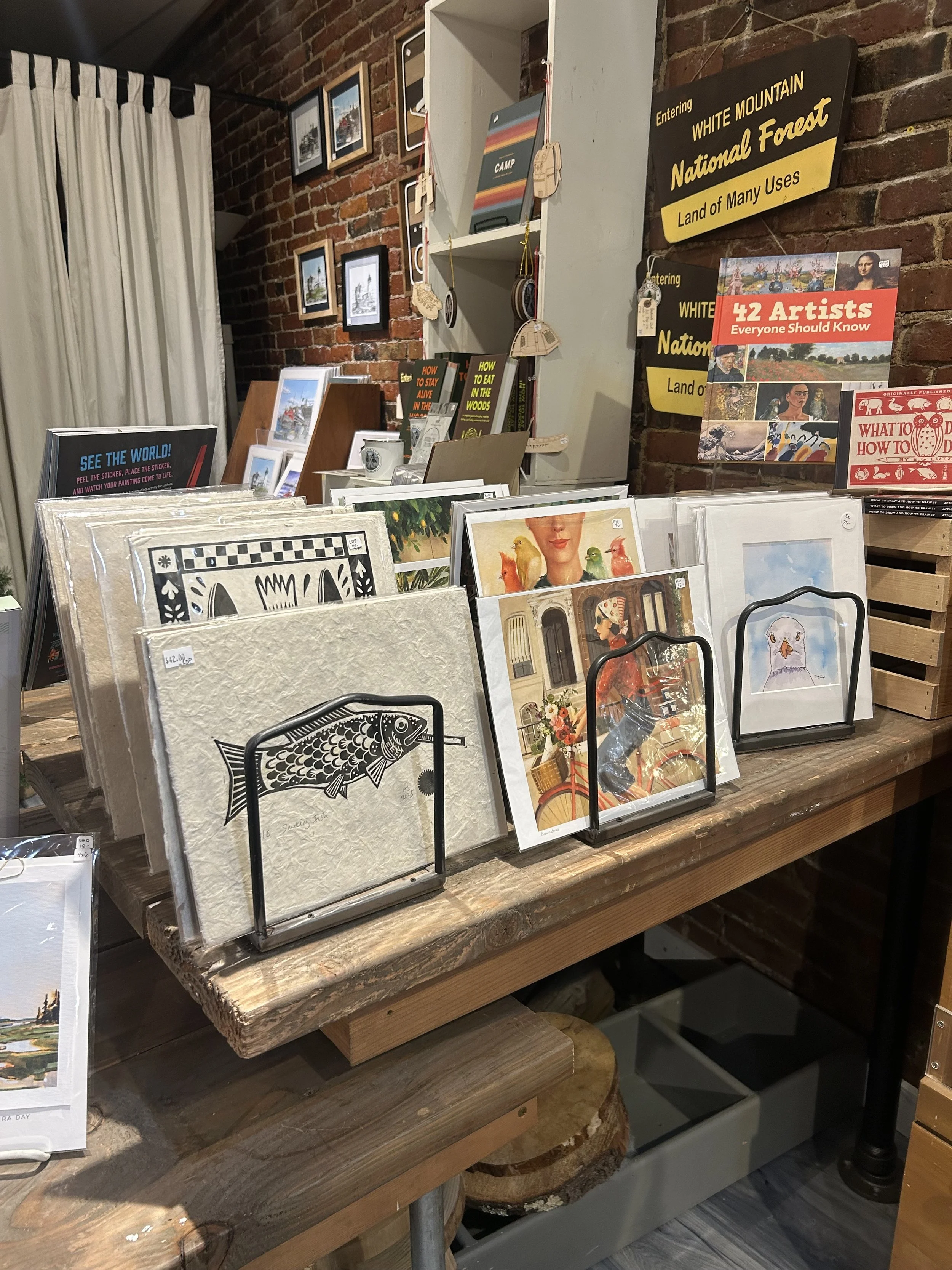 A display of framed artwork and prints on a rustic wooden table in a store, with various postcards and illustrations, including a fish, a woman on a bicycle, and other artistic images. The background features a brick wall with framed pictures and she