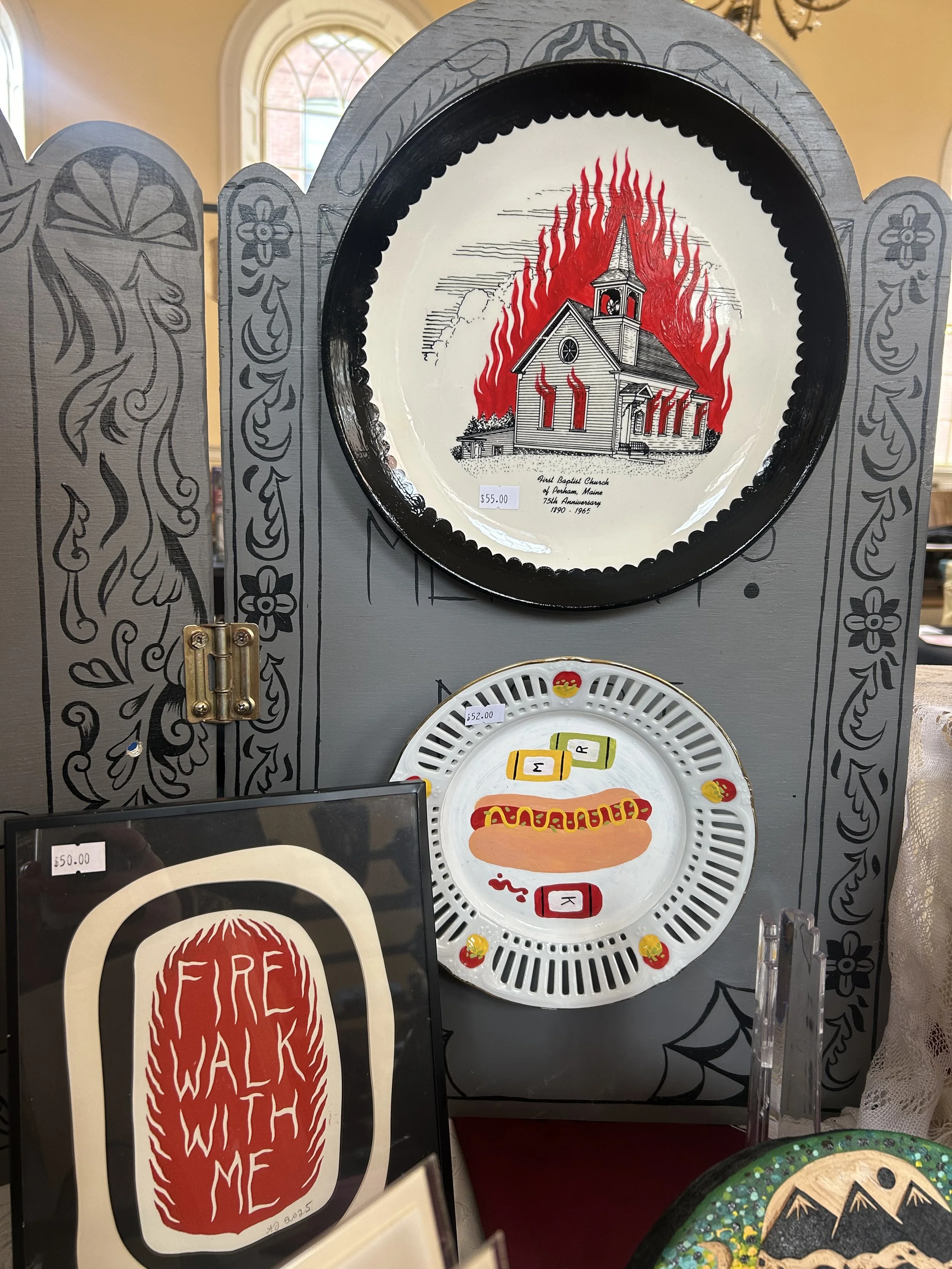 Decorative display with a round plate featuring a church on fire, a painted ceramic dish with colorful patterns, and a framed sign that reads 'Fire Walk With Me.'