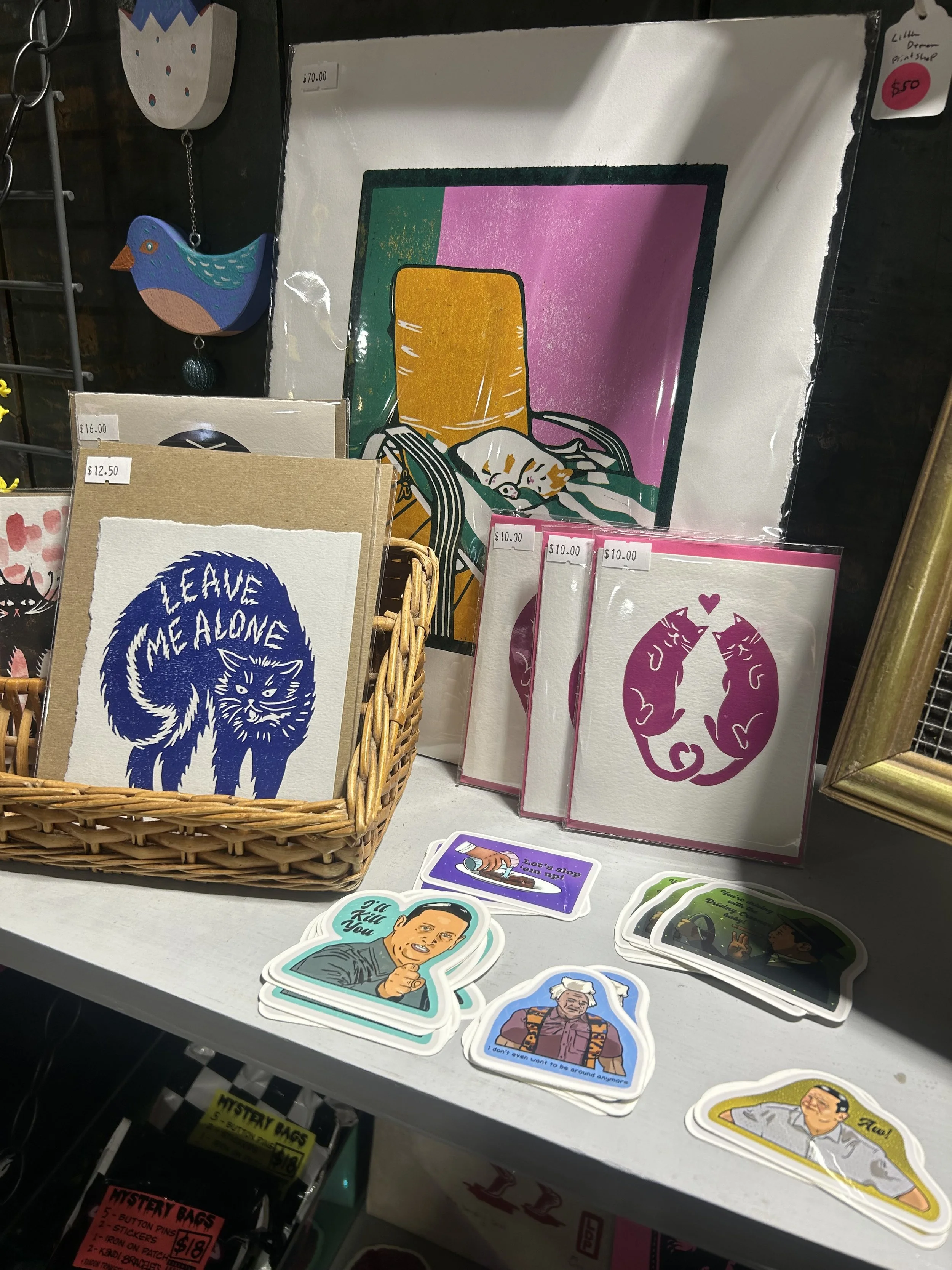 A display of various stickers and greeting cards, including a blue cat with the text Max Leave Me Alone, two pink cards with cats, and assorted illustrated stickers featuring people and humorous phrases. There is also a colorful abstract painting in 