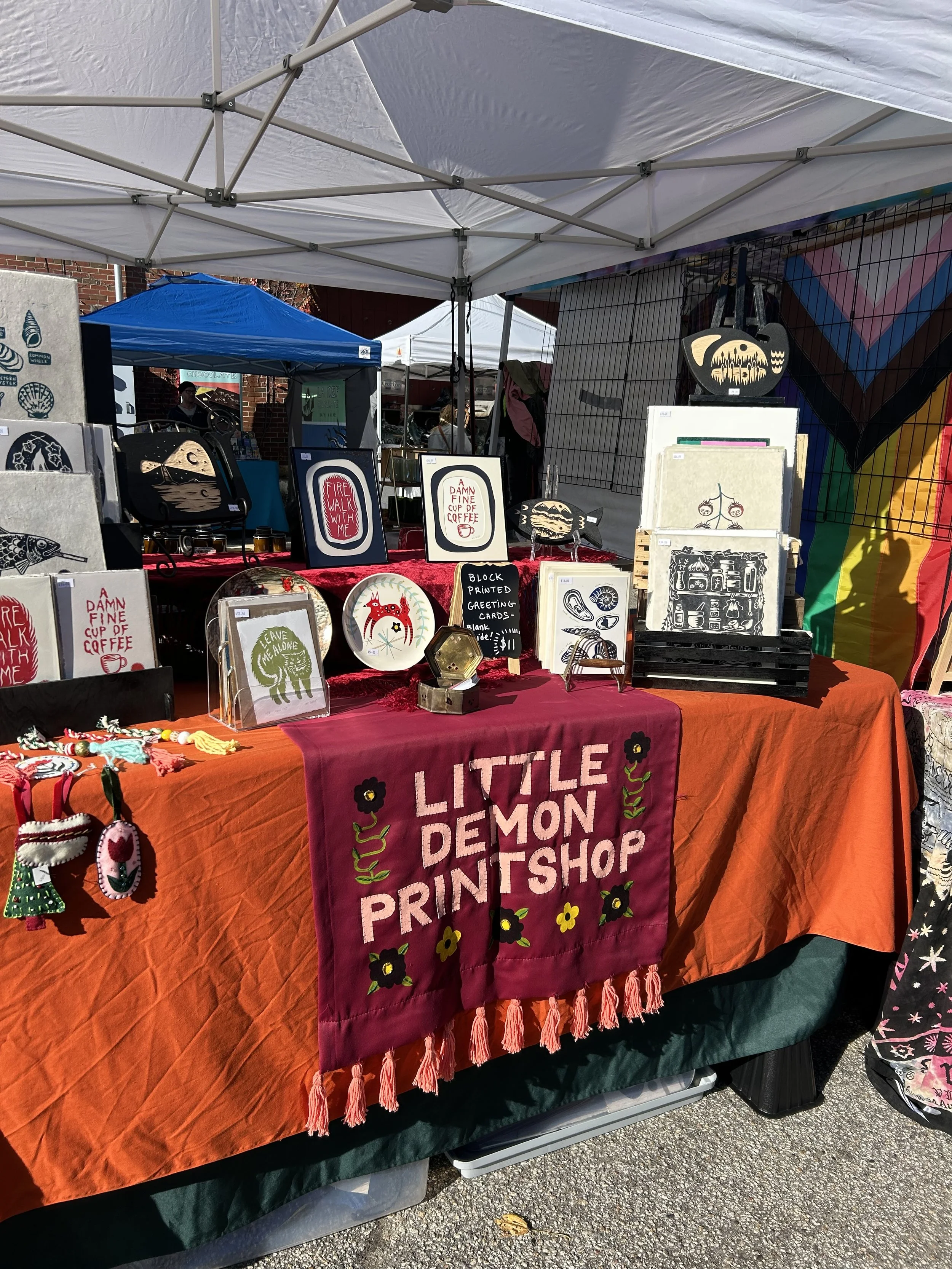 Outdoor craft stall with a red tablecloth reading "Little Demon Print Shop" displaying various printed art pieces, greeting cards, and handmade items at a market.