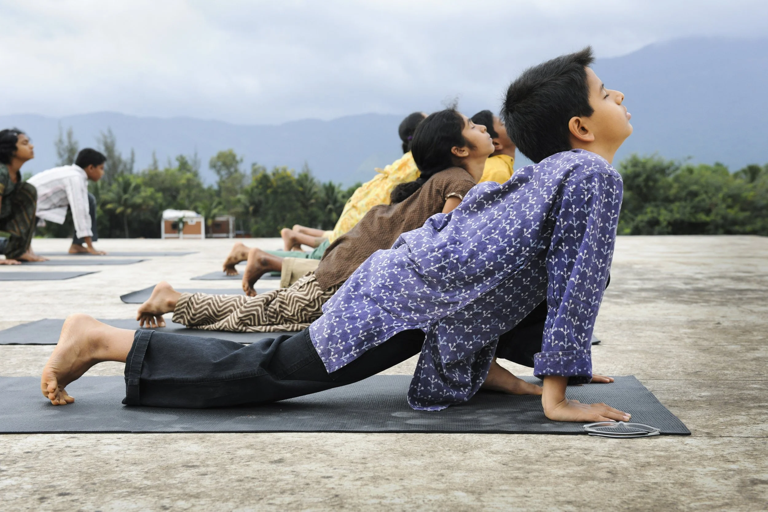 Children practicing yoga outdoors on mats, lying on their stomachs with their hands and feet on the ground, facing a scenic backdrop of mountains and trees.