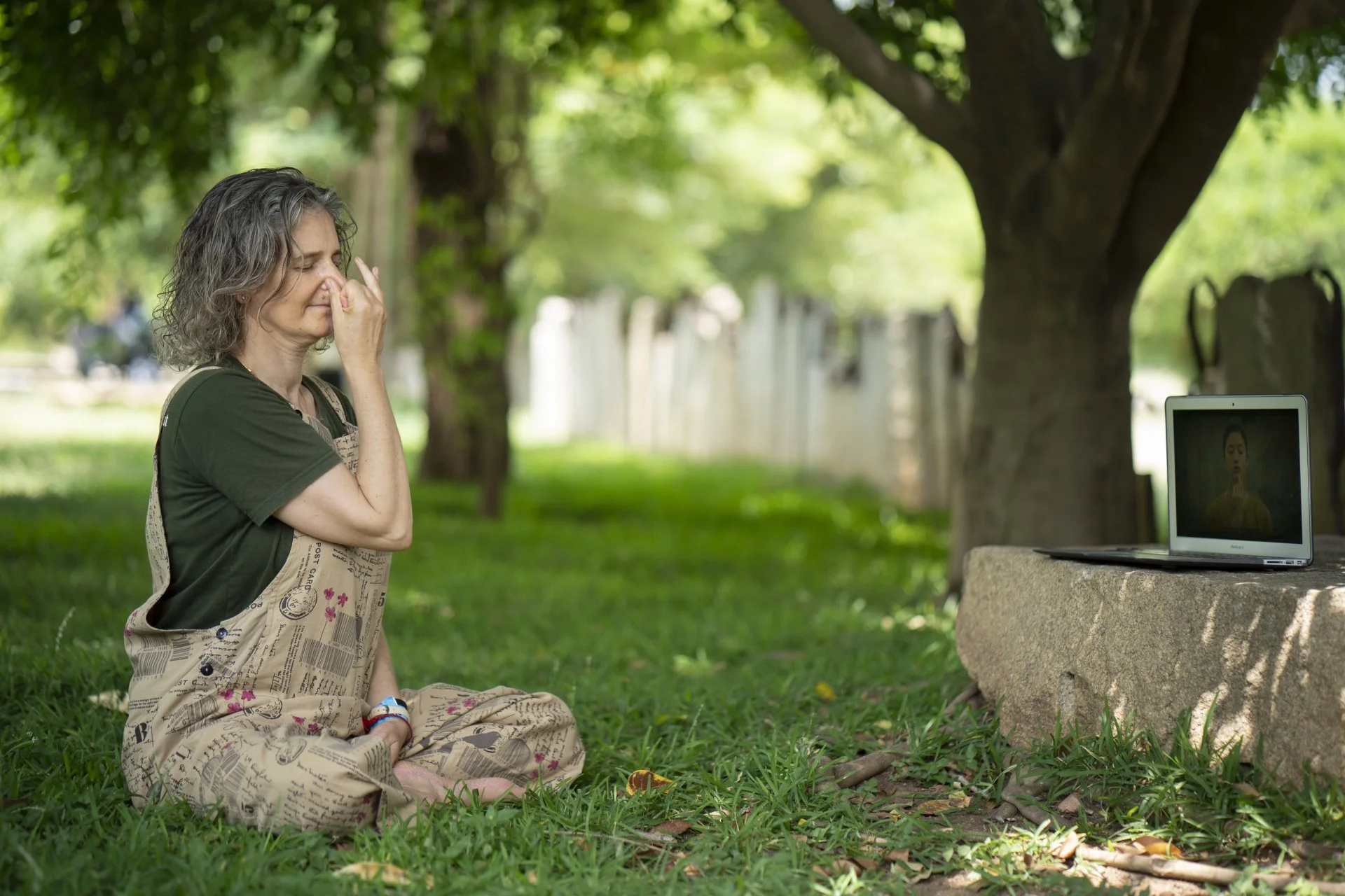 A woman with gray hair sitting cross-legged outdoors on grass, with a tree and fence in the background. She is wearing a dress with a newspaper print pattern over a dark green shirt and appears to be emotional, pinching her nose or eyes closed, facing a small laptop on a stone platform displaying a person.
