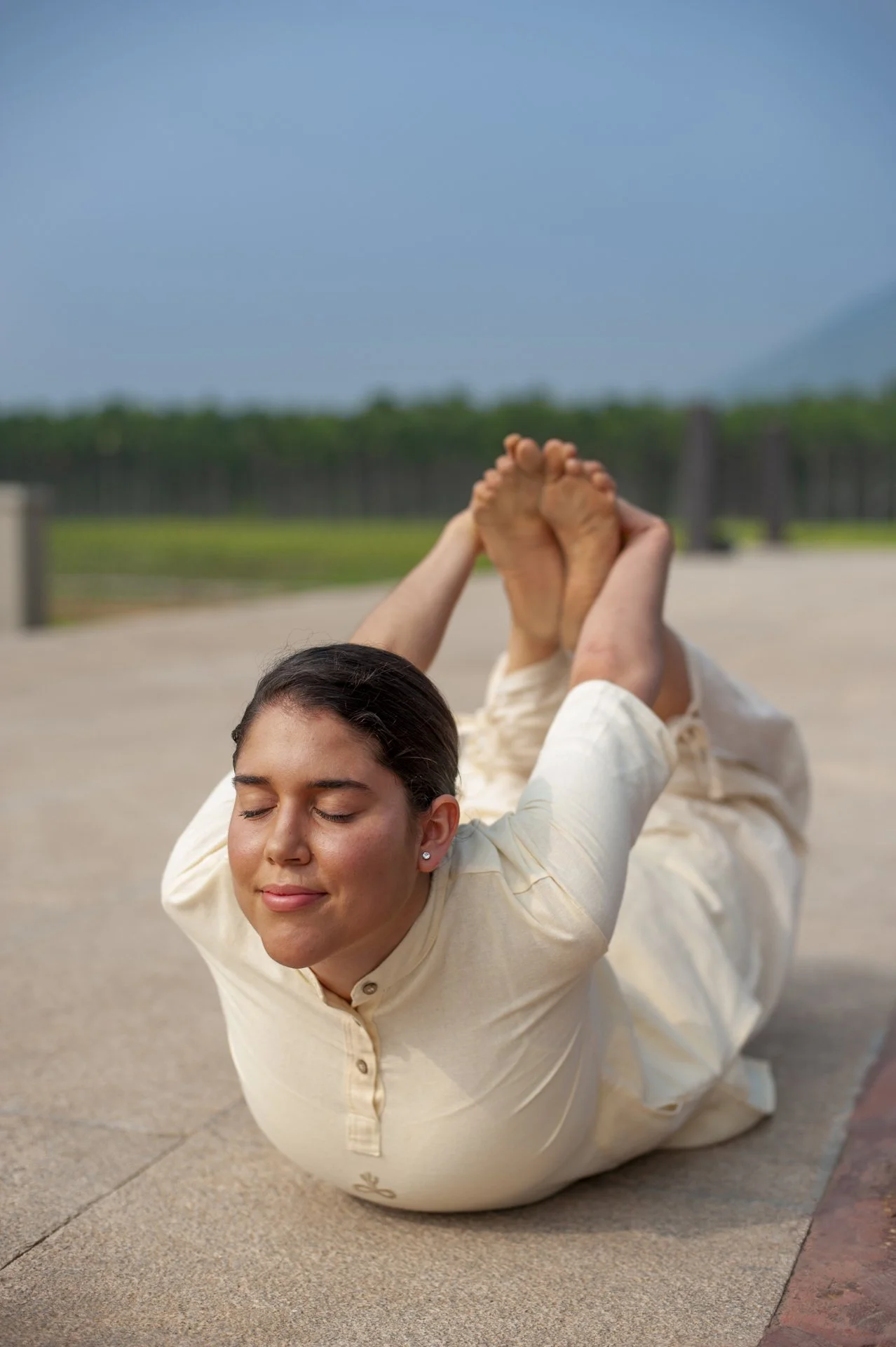 A woman practicing yoga outdoors, lying on the ground with legs bent and feet held by hands behind her head, eyes closed, and a calm expression. She is practising yogasanas a classical hatha yoga practice in Melbourne.