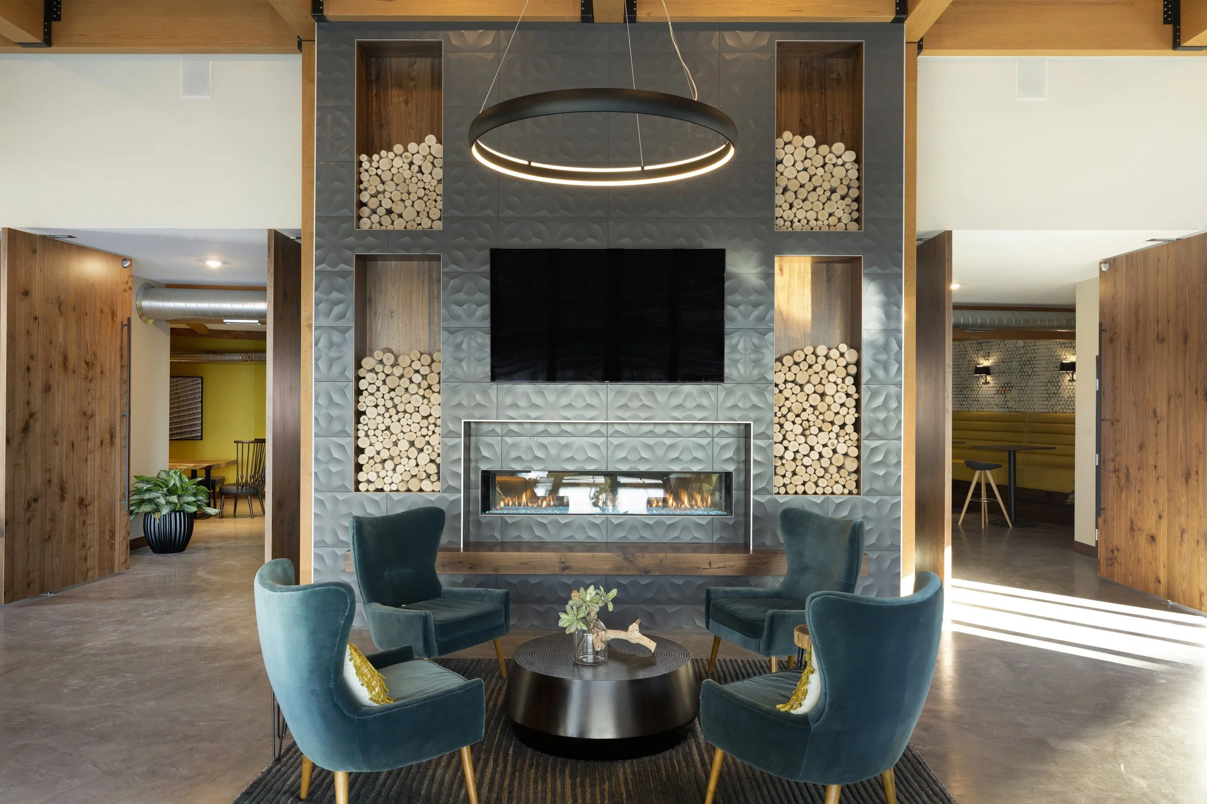 Interior of a modern lounge with four teal velvet chairs arranged in a circle around a black coffee table, a textured gray wall with a mounted flat-screen TV, a rectangular fireplace, wood accents, and decorative wood logs in built-in niches, with additional seating areas visible in the background.