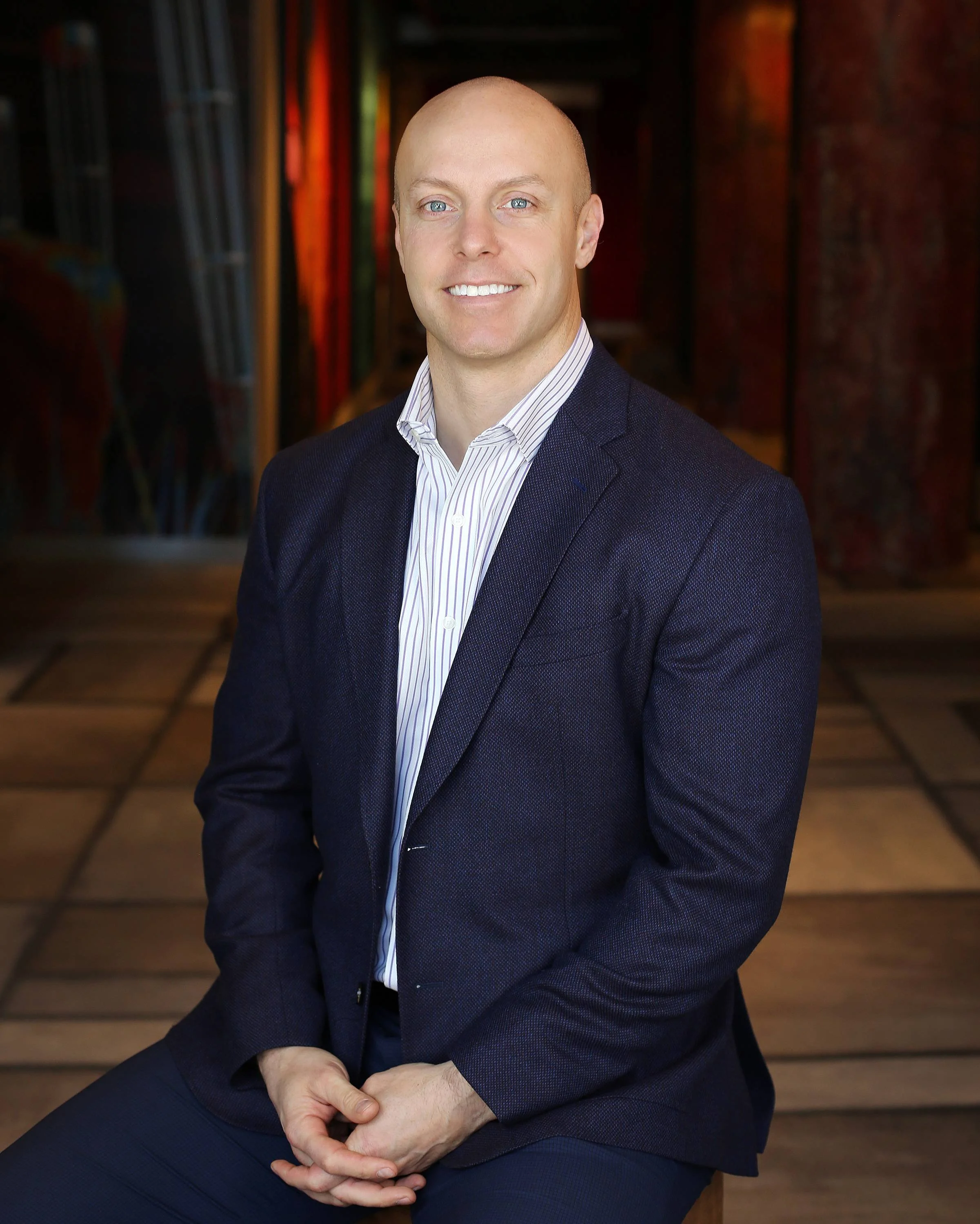 A man in a navy blazer and a striped dress shirt sitting in front of a colorful art installation background.